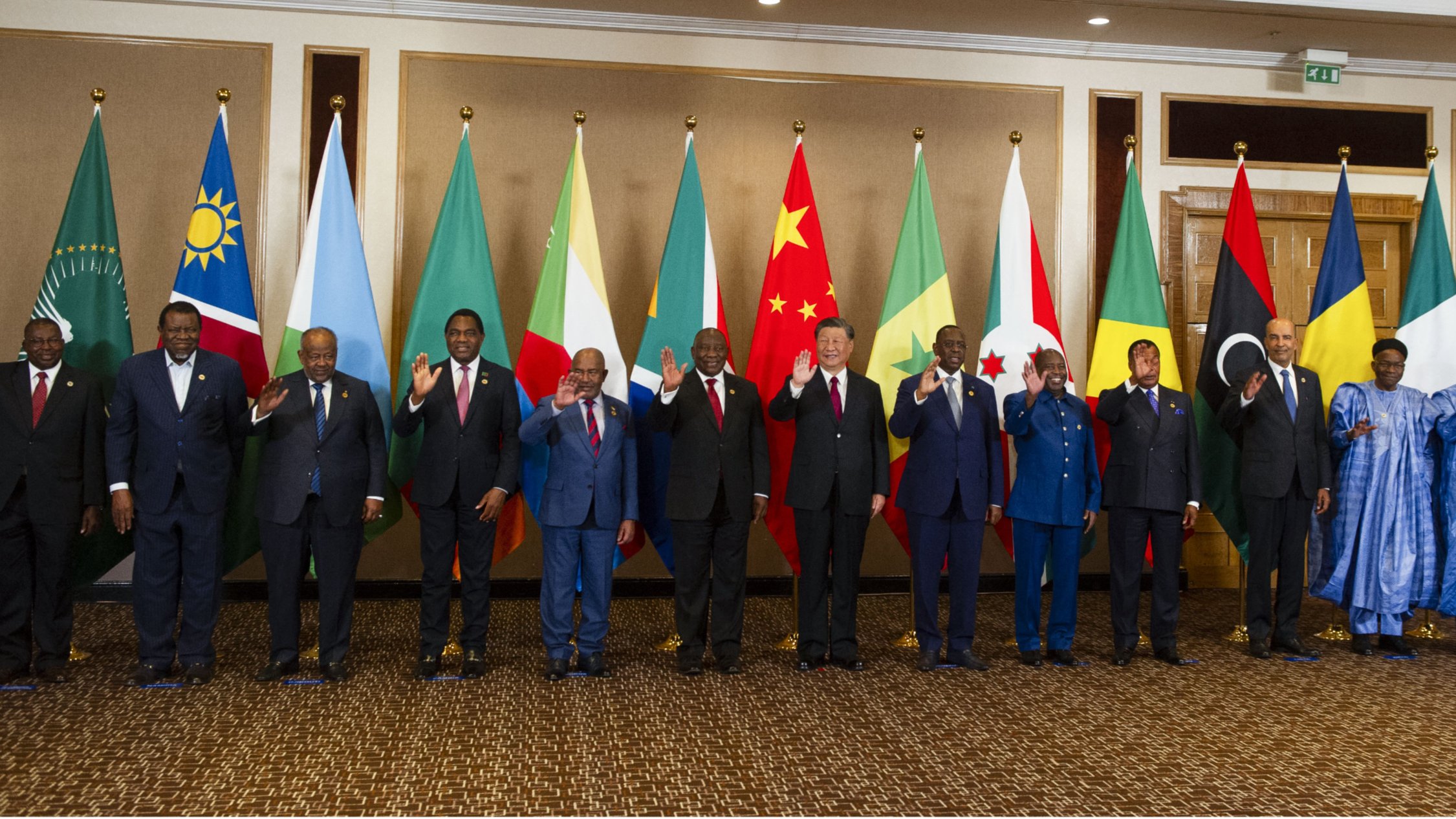 L'image montre un groupe de personnes debout en rangée, saluant. Elles se trouvent devant une série de drapeaux représentant différents pays. Les personnes portent des costumes formels et semblent participer à un événement diplomatique ou à un sommet international. Les drapeaux en arrière-plan incluent ceux de l'Union africaine, de la Chine, ainsi que d'autres nations. L'ambiance générale semble conviviale et respectueuse.
