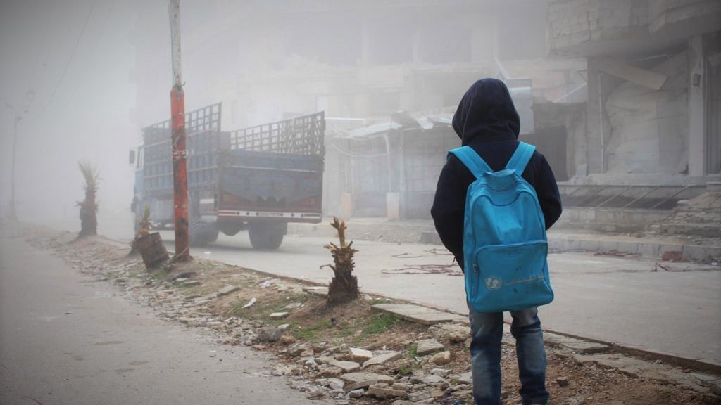 L'image montre un jeune garçon de dos, portant un grand sac à dos bleu. Il se tient sur une rue où la visibilité est réduite en raison d'un épais brouillard. En arrière-plan, on peut apercevoir des bâtiments partiellement détruits et un camion stationné. Le sol est jonché de débris et de petites plantes. L'atmosphère est sombre et mélancolique, évoquant une scène de désolation.