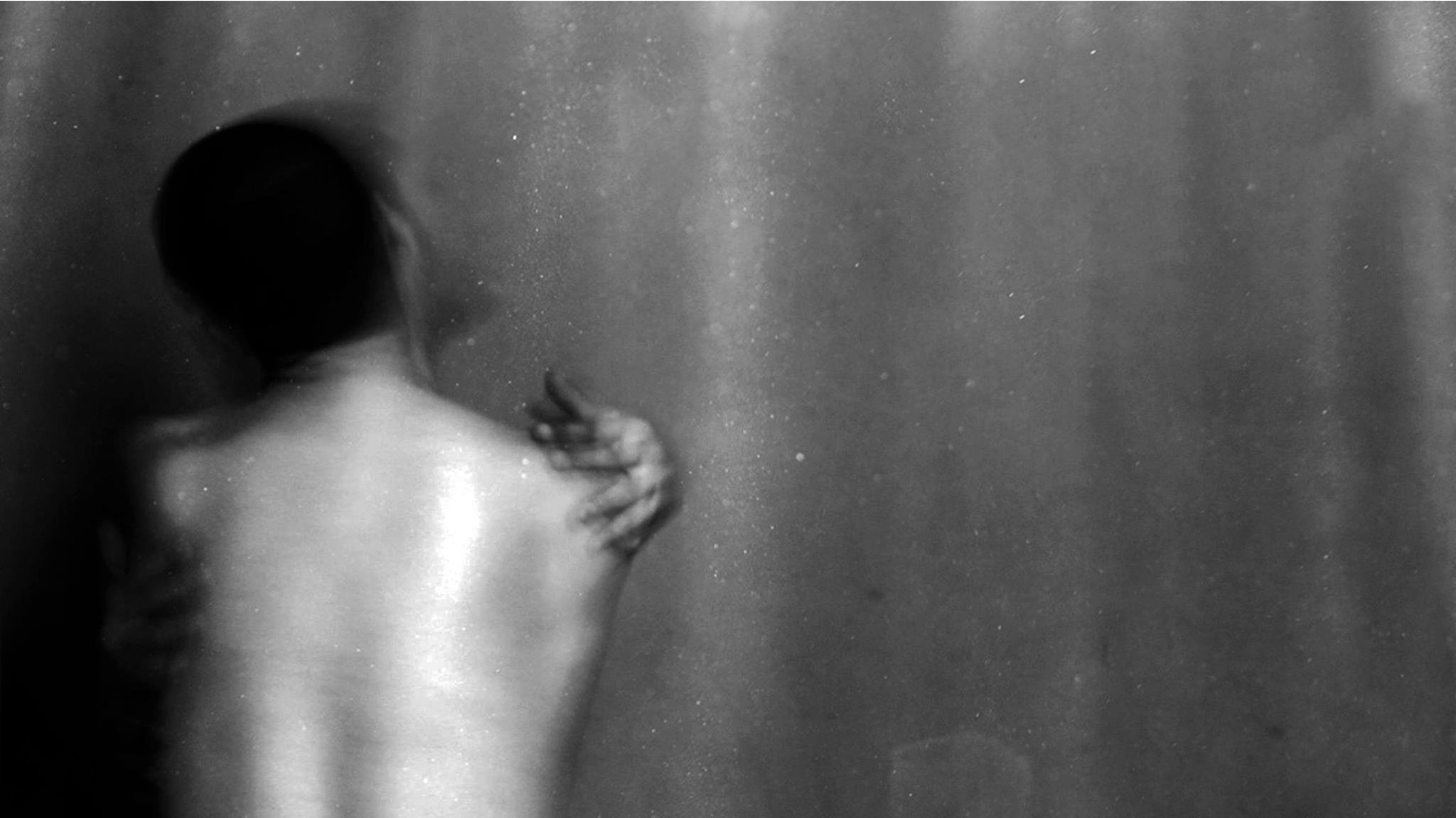 L'image montre une silhouette floue d'une personne de dos, avec un éclairage doux qui fait ressortir les contours de son corps. L'arrière-plan est sombre, créant une atmosphère intime et mystérieuse. La posture de la personne évoque une certaine vulnérabilité, tandis qu'une main semble s'accrocher à son épaule, suggérant une interaction ou un lien émotionnel. L'utilisation du noir et blanc renforce l'aspect dramatique et introspectif de la scène.