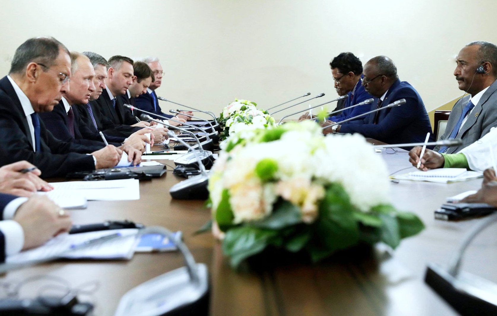 L'image représente une réunion ou une conférence avec plusieurs participants assis autour d'une grande table. Au centre de la table, il y a un arrangement floral, qui ajoute une touche de décoration. Les participants semblent être concentrés et engagés dans la discussion, prenant des notes ou écoutant attentivement. L'atmosphère est sérieuse et professionnelle, suggérant un dialogue important entre les différentes parties présentes.