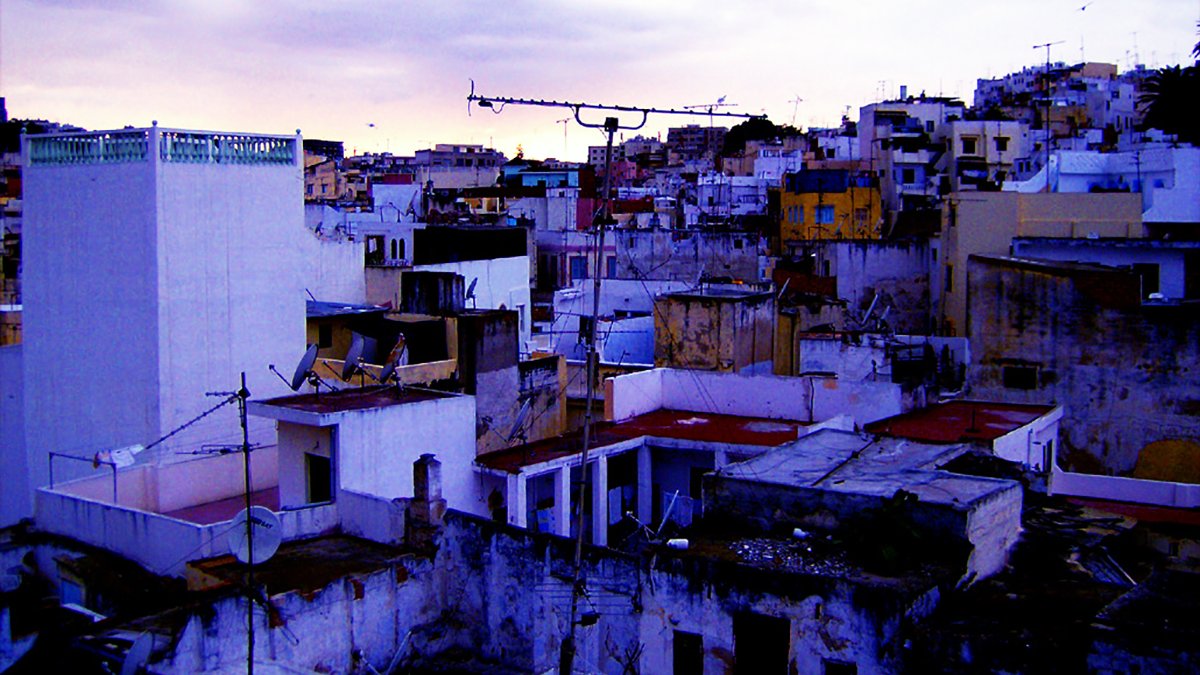 L'image montre une vue urbaine d'un paysage de ville, probablement à la tombée de la nuit. On peut voir plusieurs bâtiments aux structures variées, avec des toits plats et un mélange de couleurs qui évoquent une atmosphère un peu mélancolique. Certains bâtiments semblent légèrement en ruine, avec des murs décrépis et des débris visibles. Le ciel est nuageux, ajoutant une ambiance dramatique à la scène. Des antennes de télévision sont visibles sur les toits, typiques des environnements urbains. L'ensemble donne une impression de vie quotidienne dans une ville qui a du caractère.