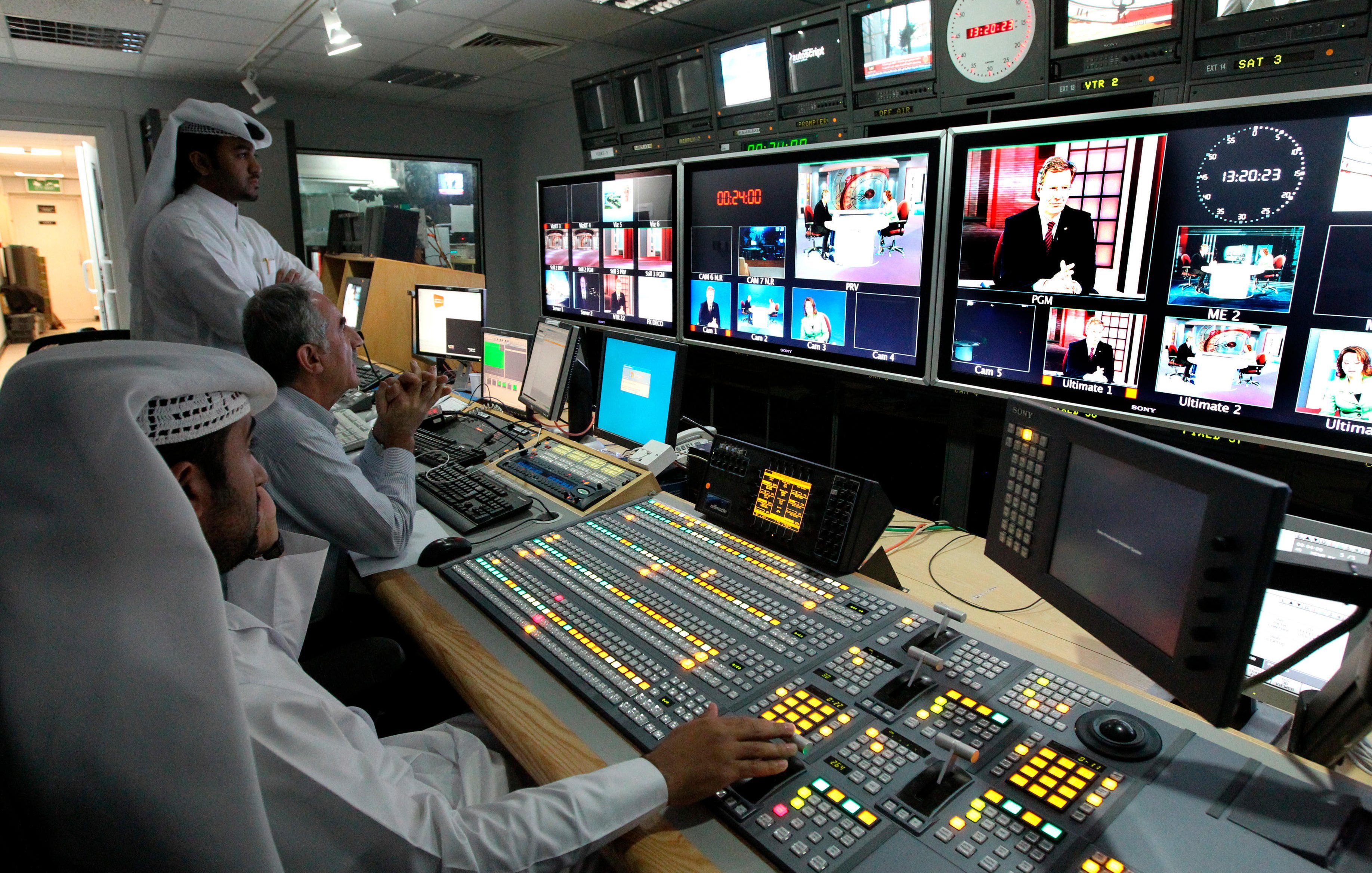 L'image montre un intérieur d'une salle de contrôle de diffusion. On y voit plusieurs personnes travaillant sur des panneaux de contrôle avec des écrans affichant différentes émissions. Au premier plan, une personne est assise devant une console, mesurant les niveaux et ajustant les paramètres. Deux autres personnes se tiennent derrière, observant attentivement les écrans. Les murs sont équipés de plusieurs moniteurs affichant des vidéos en direct ou des reportages. L'ambiance semble concentrée et professionnelle, typique d'un environnement de télévision.