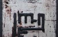 L'image montre une œuvre d'art abstraite sur un fond blanc texturé, avec des traces de rouge qui semblent représenter des coups de pinceau ou des éclaboussures. Au centre, il y a des lignes noires qui forment un motif géométrique, évoquant un symbole ou une écriture stylisée. Le cadre est en bois noir, ajoutant un contraste à l'œuvre. L'ensemble dégage une atmosphère unique et intrigante, mélangeant des éléments de peinture et de sculpture.