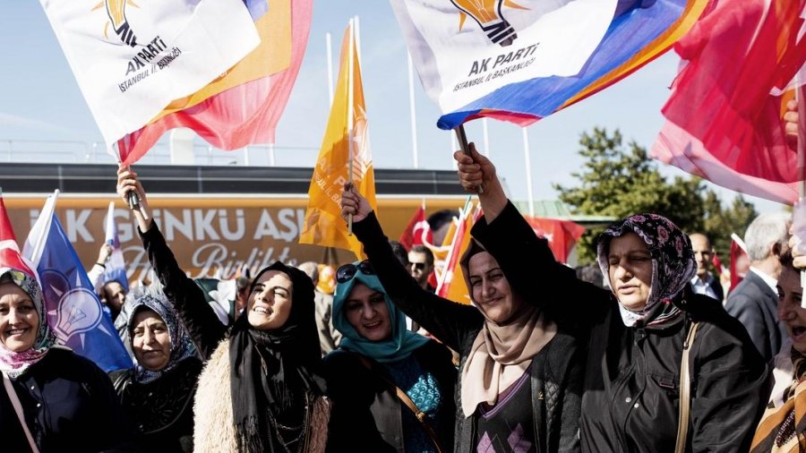L'image montre un groupe de femmes rassemblées lors d'un événement ou d'une manifestation, brandissant des drapeaux. Ces drapeaux portent le logo d'un parti politique, probablement le Parti de la justice et du développement (AK Parti) en Turquie. Les femmes semblent enthousiastes et engagées, portant des vêtements variés, y compris des foulards. L'arrière-plan présente d'autres drapeaux et des affiches, créant une ambiance festive et militante.