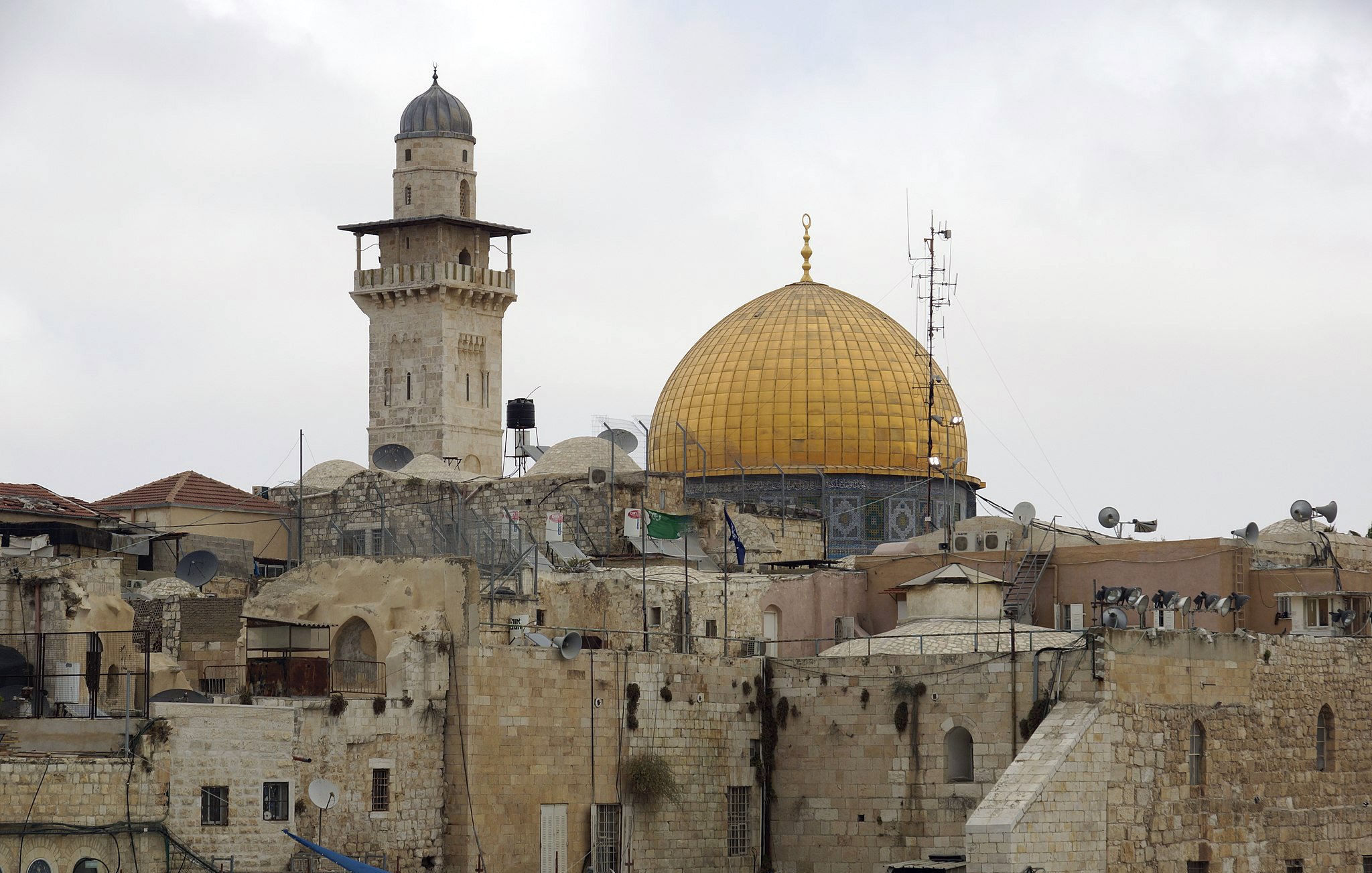 L'image montre une vue d'un paysage urbain avec des bâtiments en pierre. Au centre, on peut apercevoir un dôme doré, probablement celui du Rocher, qui est un site religieux important. À proximité, on remarque un minaret qui surplombe les structures anciennes. Le ciel est nuageux, ajoutant une atmosphère douce à la scène. Les toits sont parsemés de paraboles satellites, témoignant de la modernité mêlée à l'histoire de cette région.