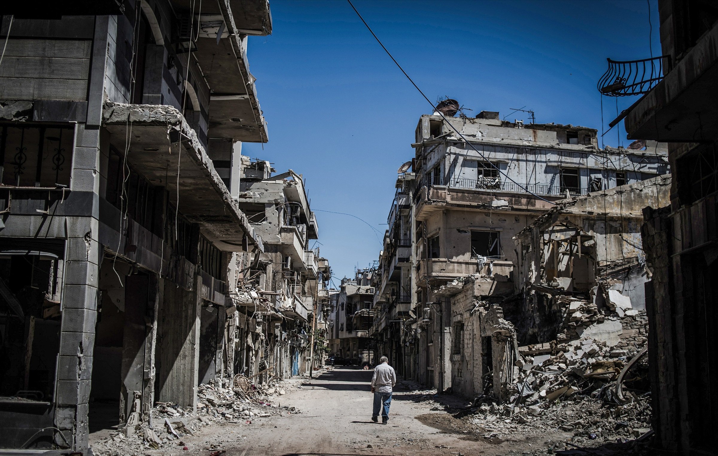 L'image montre une rue dévastée, bordée de bâtiments en ruine. Les murs sont fissurés et effondrés, témoignant des destructions causées par un conflit. Au centre de la scène, un homme marche seul, semblant contempler les vestiges de ce qui était autrefois un quartier vivant. Le ciel est clair, contrastant avec la désolation environnante. Cette image évoque une atmosphère de solitude et de tristesse face à la perte et à la destruction.