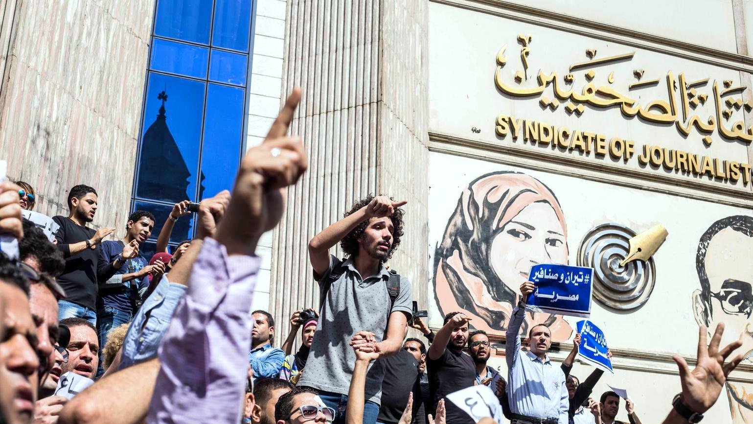L'image montre une scène de manifestation devant un bâtiment, probablement le Syndicat des journalistes. De nombreuses personnes sont rassemblées, levant les bras en signe de protestation. Certains tiennent des pancartes ou des banderoles, tandis qu'une personne debout sur une foule semble s'adresser aux manifestants. L'arrière-plan présente une grande fresque représentant des figures, ajoutant à l'impact visuel de la scène. L'atmosphère est celle d'un rassemblement engagé et passionné.