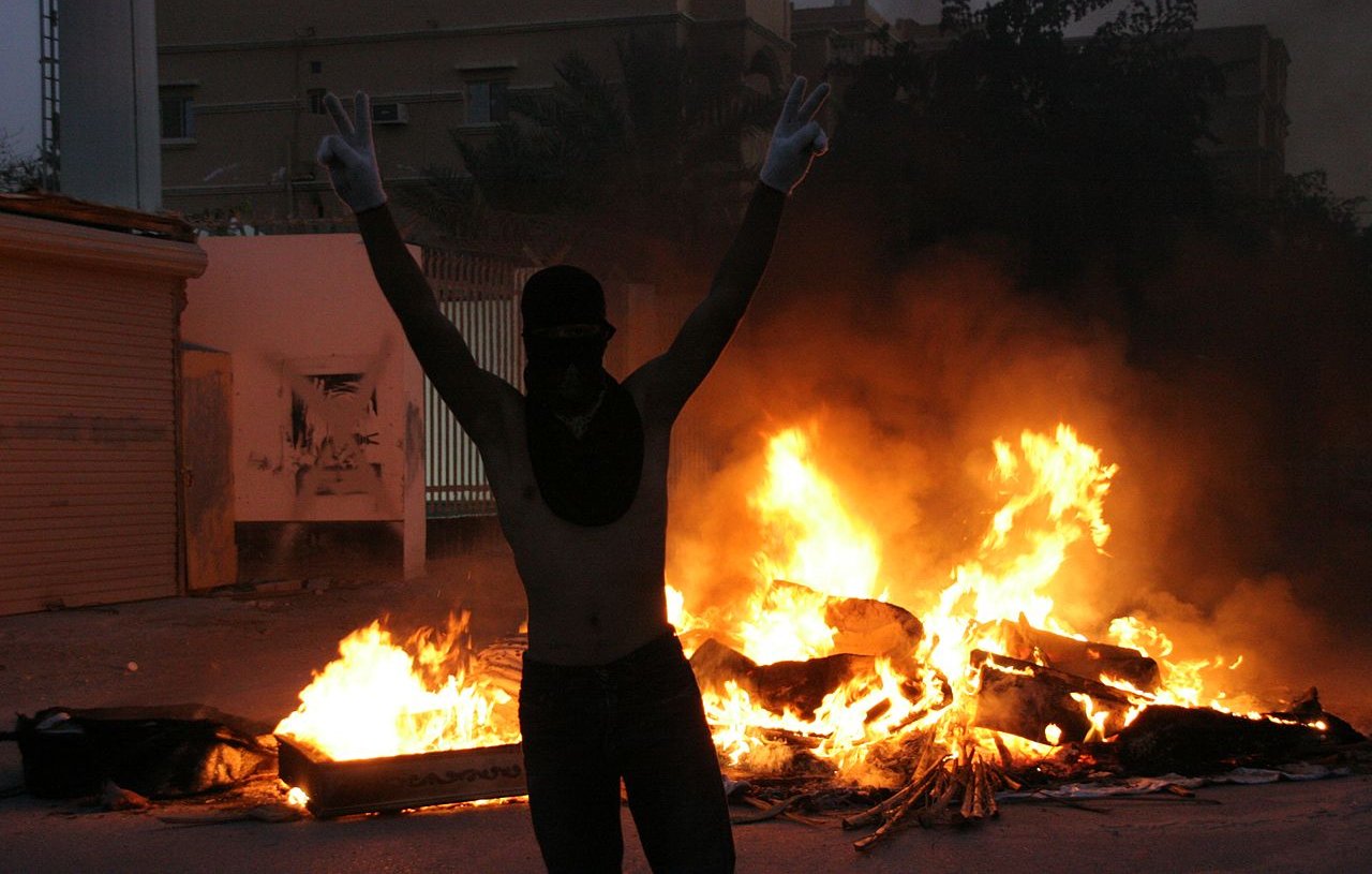 L'image montre une scène intense et dramatique. À l'avant-plan, une personne se tient avec les bras levés, formant un signe de victoire, tout en portant un masque qui couvre son visage. En arrière-plan, on peut voir des flammes vives qui s'élèvent, indiquant un feu, probablement une barricade ou des objets en feu. L'atmosphère est chargée d'émotion, évoquant un moment de protestation ou de turbulence. Les ombres des bâtiments environnants ajoutent à l'ambiance sombre de la scène.