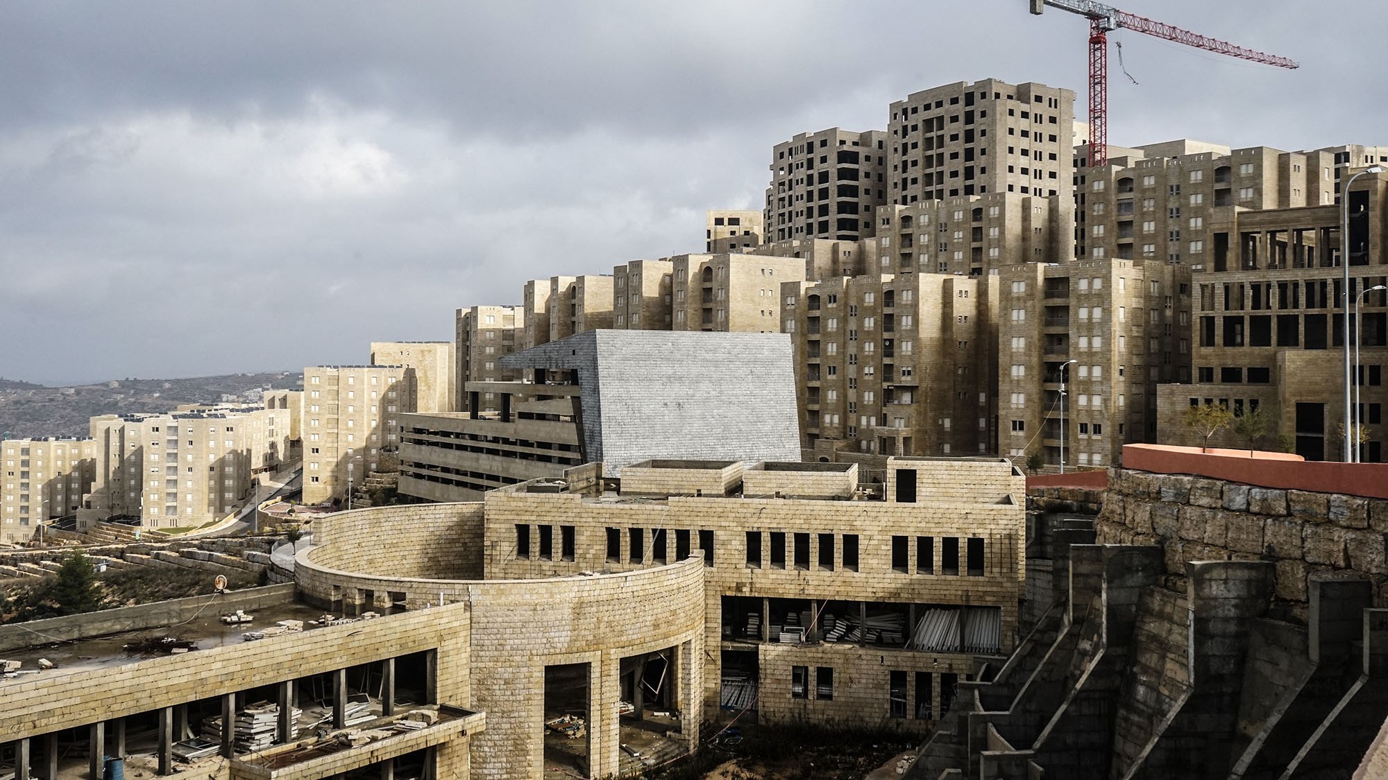 L'image montre un paysage urbain en construction, avec des bâtiments modernes de forme géométrique et des structures encore inachevées. On peut voir des façades en béton et des fenêtres non terminées sur plusieurs des bâtiments. En arrière-plan, une grue rouge indique que des travaux sont en cours. Le ciel est nuageux, créant une atmosphère un peu sombre. Au premier plan, des éléments de construction sont visibles, suggestifs d'un site en développement.