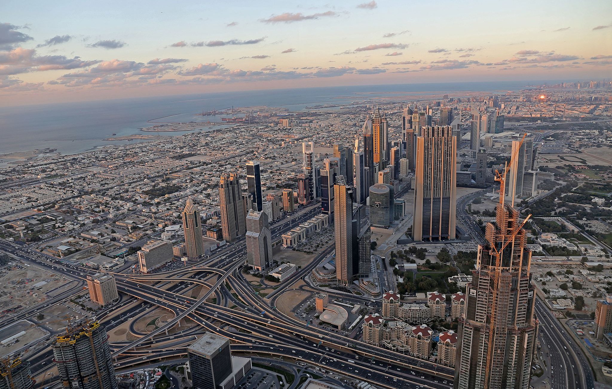 L'image montre un panorama impressionnant d'une ville moderne, probablement Dubai. On y voit de nombreux gratte-ciels élégants s'élevant vers le ciel, ainsi qu'un réseau complexe de routes et d'autoroutes serpentant à travers la métropole. À l'arrière-plan, on aperçoit la mer et des paysages urbains qui s'étendent à perte de vue. Le ciel présente des teintes douces de bleu et d'orange, témoignant d'un coucher de soleil, ce qui ajoute une ambiance tranquille à la scène urbaine animée.
