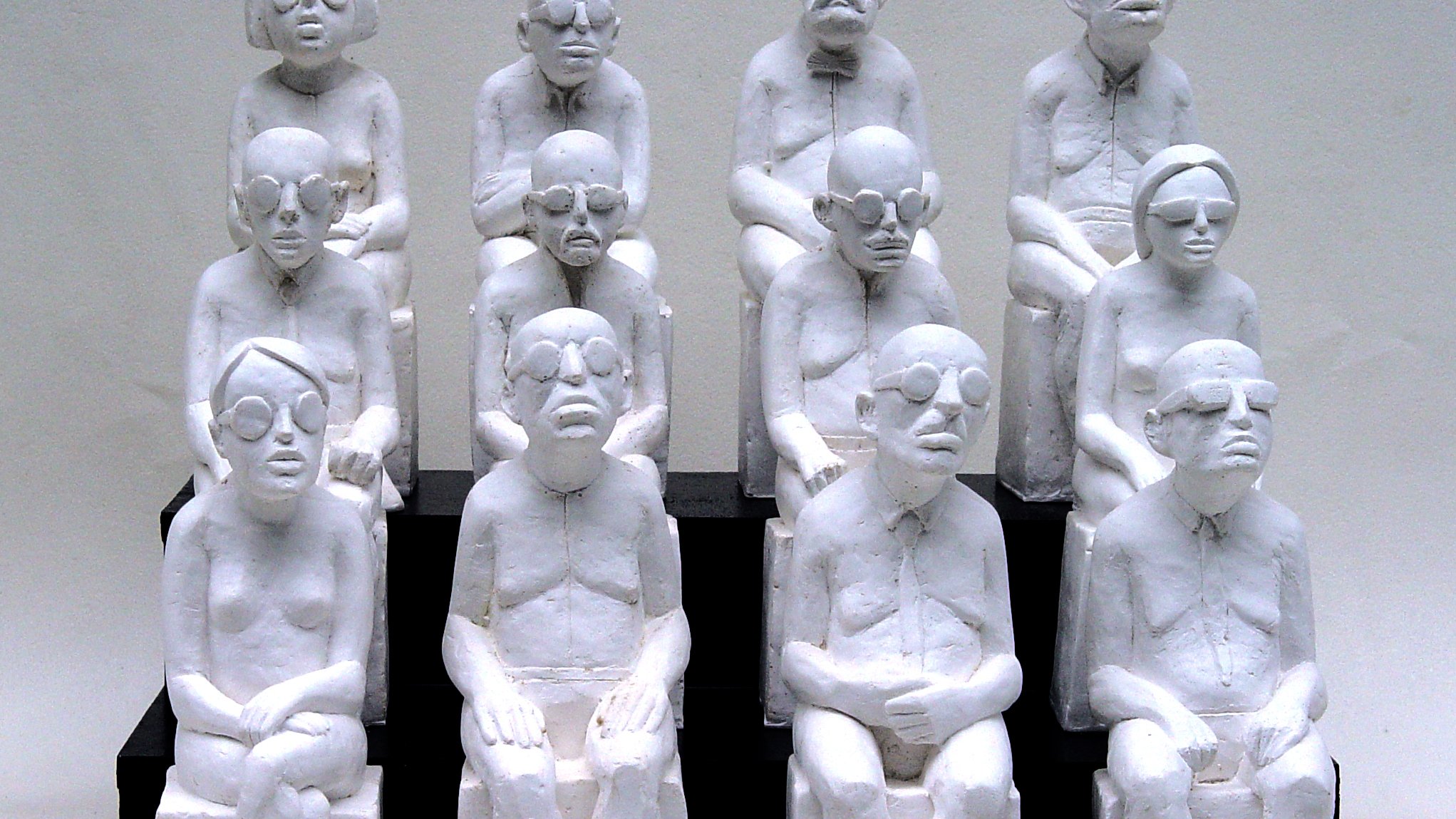L'image représente une sculpture où plusieurs figures humaines, toutes de couleur blanche, sont assises sur des bancs. Les personnages sont stylisés avec des traits simples et géométriques, portant des lunettes, et semblent avoir des expressions neutres ou pensive. Ils sont agencés en un groupe de dix, disposés en plusieurs rangées, créant une impression d'unité et de contemplation. Les sculptures semblent évoquer des thèmes tels que la solitude, la réflexion ou la condition humaine.