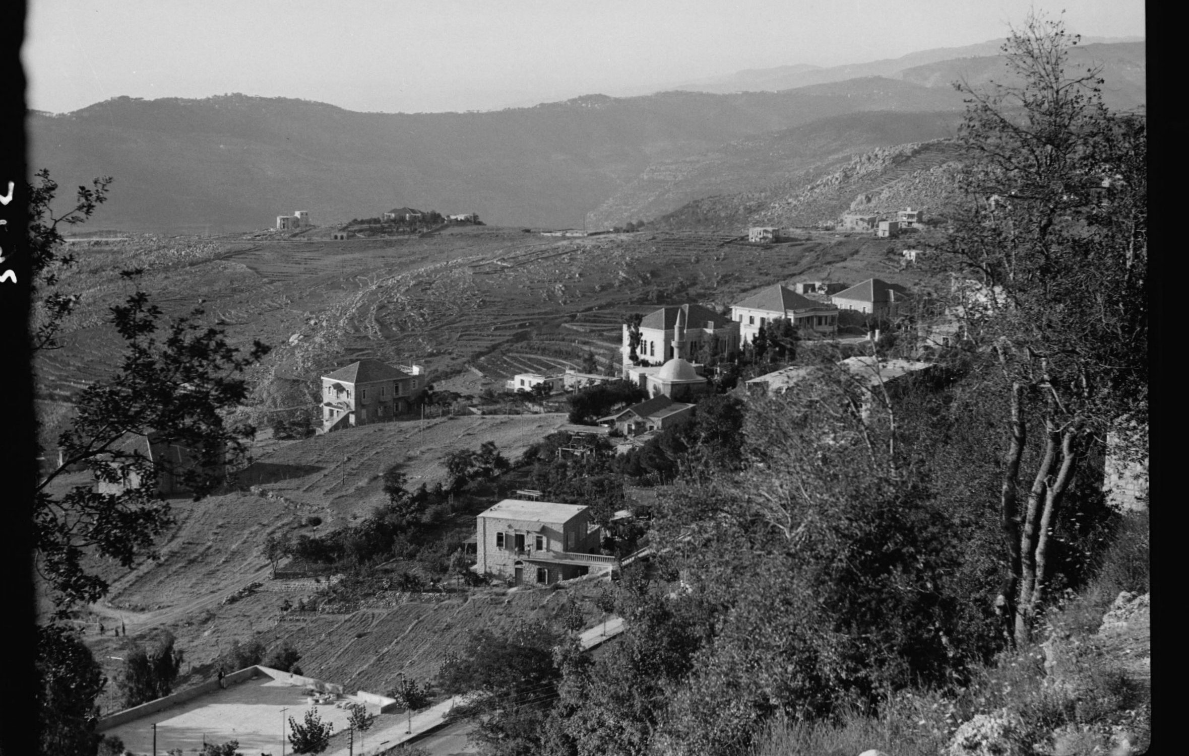 L'image montre un paysage montagneux avec des collines qui s'étendent à l'horizon. On peut apercevoir plusieurs maisons éparpillées sur les flancs des collines, entourées de végétation. La scène semble être prise en noir et blanc, ce qui lui donne un aspect nostalgique. Les terrains agricoles sont visibles, indiquant une zone rurale. Les montagnes en arrière-plan ajoutent une profondeur à la composition paysagère.