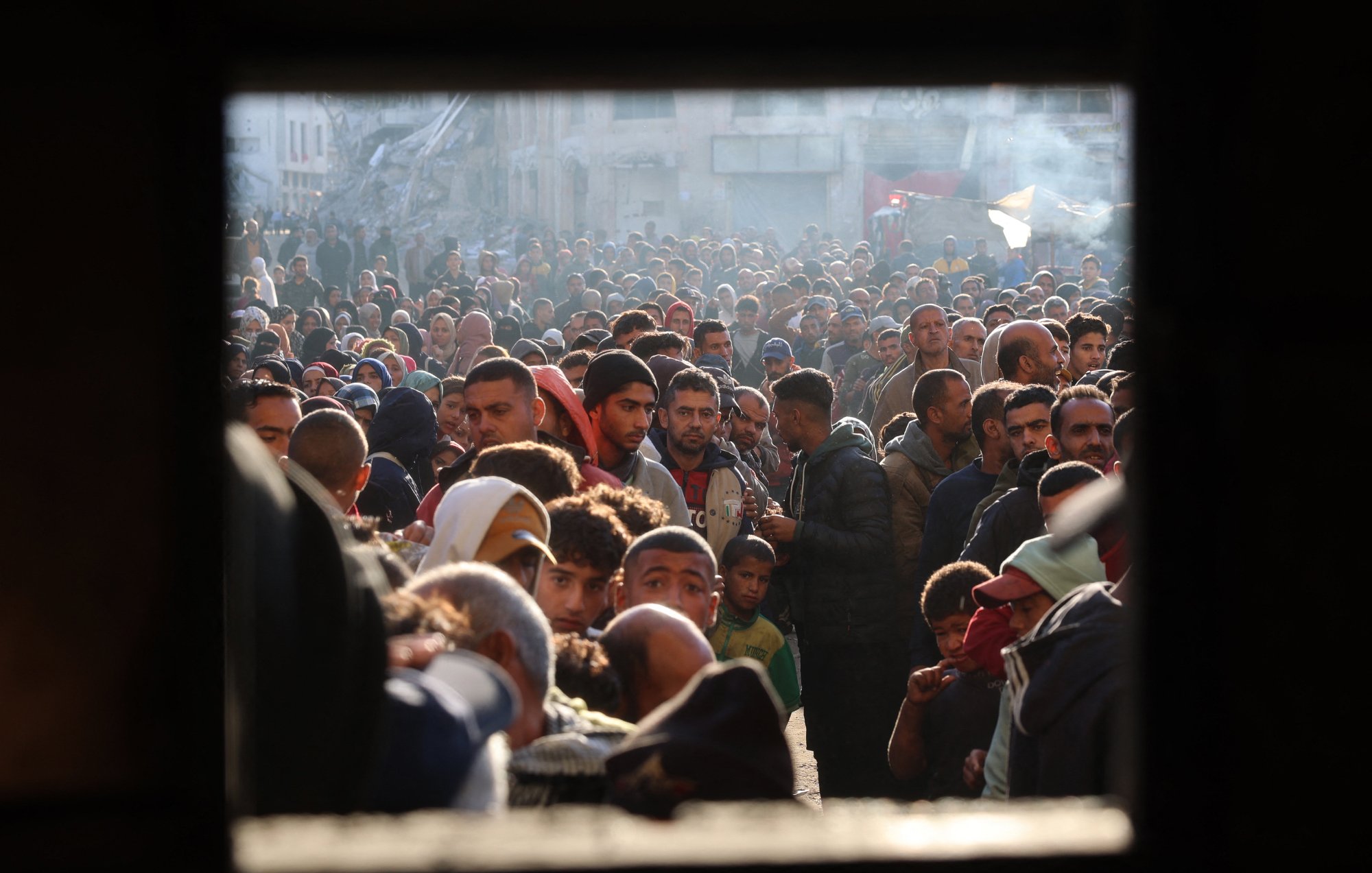 L'image montre une foule dense de personnes, probablement rassemblées dans un espace urbain. Les visages sont variés, et l'atmosphère semble chargée d'émotion et de tension. On peut voir des hommes, des femmes et des enfants, témoignant d'une situation de forte affluence, peut-être liée à un événement particulier comme une distribution d'aide ou une manifestation. Les lumières et l'arrière-plan laissent suggérer un contexte urbain avec des bâtiments visibles. L'image est encadrée par une ouverture, ce qui donne l'impression d'un regard extérieur sur ce rassemblement.