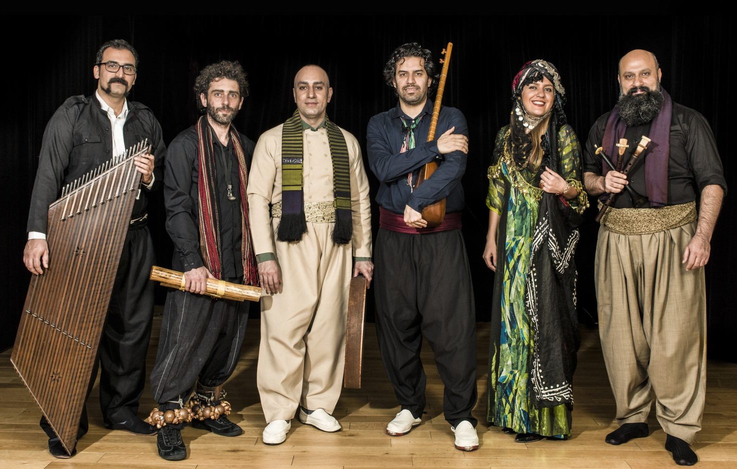 L'image montre un groupe de six musiciens posant ensemble sur scène. Ils sont vêtus de vêtements traditionnels, ce qui évoque une ambiance culturelle riche. Chaque membre tient un instrument différent : on peut voir un instrument à cordes, des percussions et d'autres instruments typiques. Leur expression est joyeuse et conviviale, et l'éclairage met en valeur leurs costumes. L'arrière-plan est sombre, ce qui fait ressortir leurs tenues colorées et détaillées. Cela donne une impression de performance musicale authentique et festive.