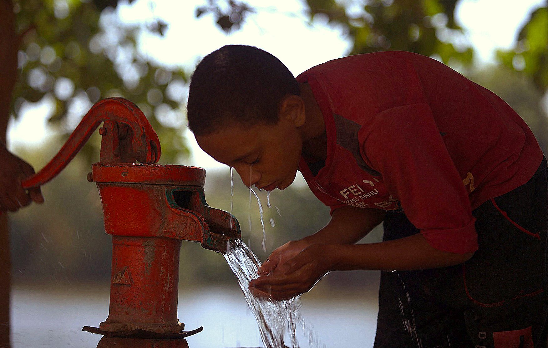 Sur l'image, un jeune garçon penche sa tête en avant pour recueillir de l'eau qui jaillit d'une pompe manuelle rouge. Il a l'air concentré et utilise ses mains pour attraper l'eau qui s'écoule. En arrière-plan, on peut apercevoir un paysage flou avec un plan d'eau. La lumière semble douce, et l'atmosphère évoque un moment de fraîcheur et de vie quotidienne.