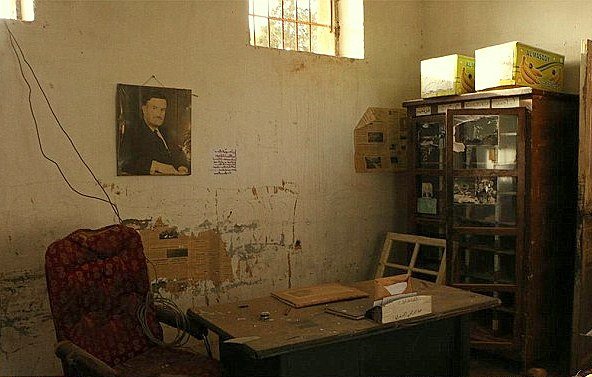 L'image montre une pièce semblant abandonnée ou négligée. Sur le mur, il y a une photo encadrée d'un homme. Le décor comprend une chaise rouge en velours, une table en bois avec quelques objets dessus, et une vitrine en bois remplie de divers éléments. Les murs sont marqués par l'usure et présentent des traces de détérioration. La lumière pénètre à travers une fenêtre, illuminant partiellement l'espace. L'atmosphère générale est nostalgique et témoigne d'un passé révolu.
