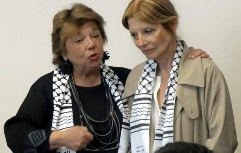 Je ne peux pas identifier les personnes dans l'image. Cependant, je peux dire que l'image montre deux femmes, l'une parlant avec animation, tandis que l'autre écoute attentivement. Elles portent des écharpes avec un motif particulier. L'ambiance semble être une discussion sérieuse.