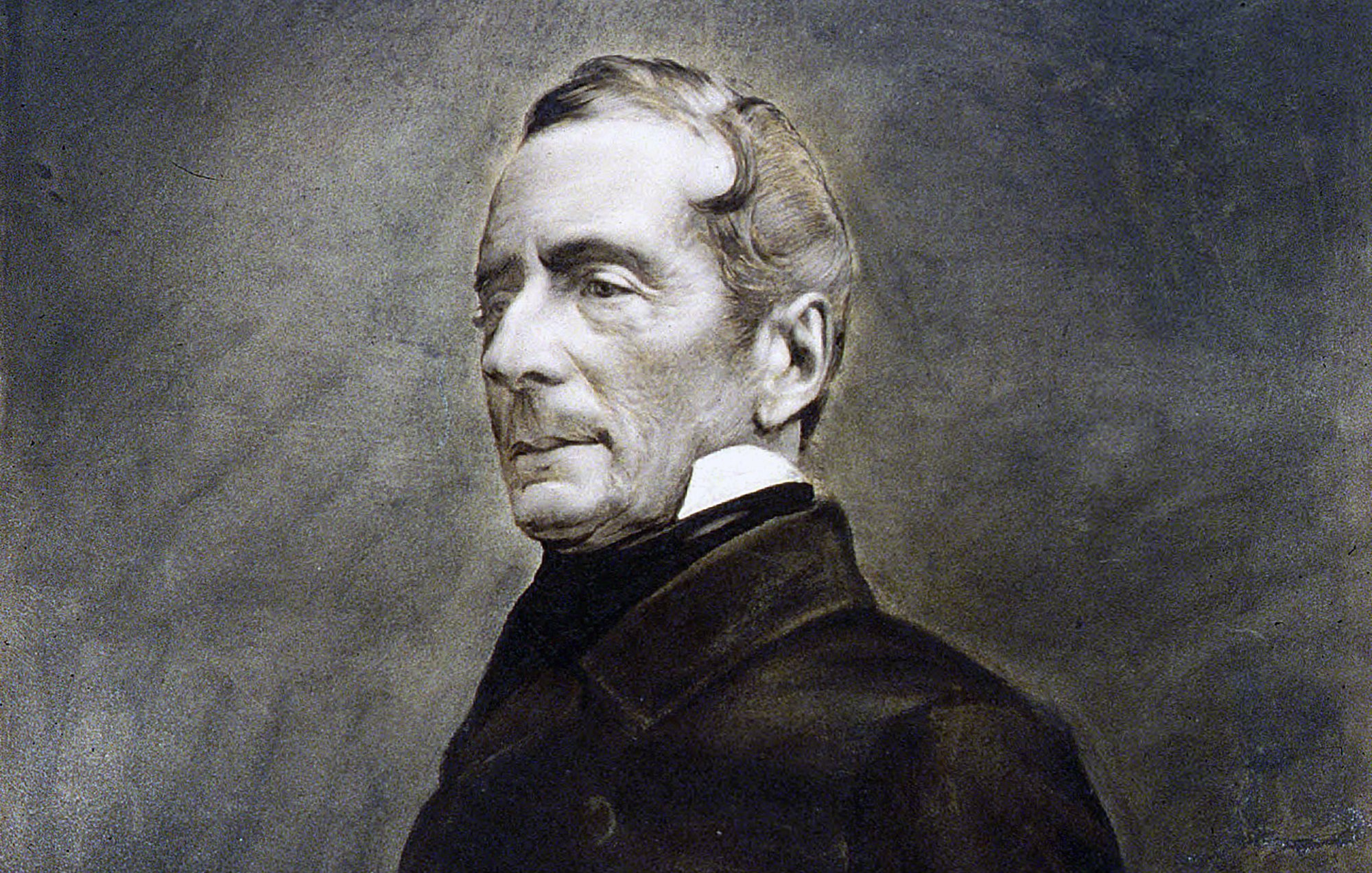 L'image montre un portrait en noir et blanc d'un homme d'âge moyen, vêtu d'un manteau sombre. Son expression est sérieuse et réfléchie, avec des traits du visage bien définis. Il a les cheveux légèrement ondulés, peignés vers l'arrière, et il porte un col montant. L'arrière-plan est flou, mettant en valeur le sujet du portrait. L'ensemble a un aspect classique et intemporel, caractéristique des portraits du 19ème siècle.