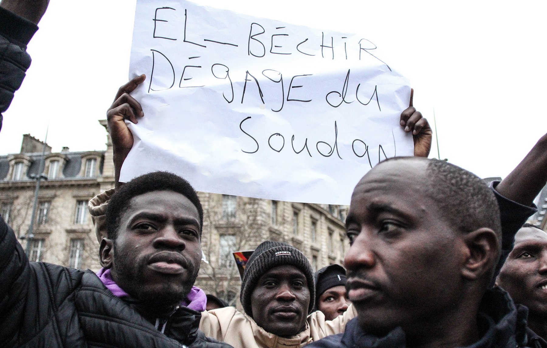 L'image montre une manifestation où des personnes tiennent des pancartes. L'une d'elles est clairement visible et affiche un message en français disant "El-Béchir dégage du Soudan". Les participants semblent passionnés et engagés dans une cause. On peut discerner des expressions sérieuses et déterminées sur leurs visages. En arrière-plan, des bâtiments peuvent être aperçus, suggérant un cadre urbain.