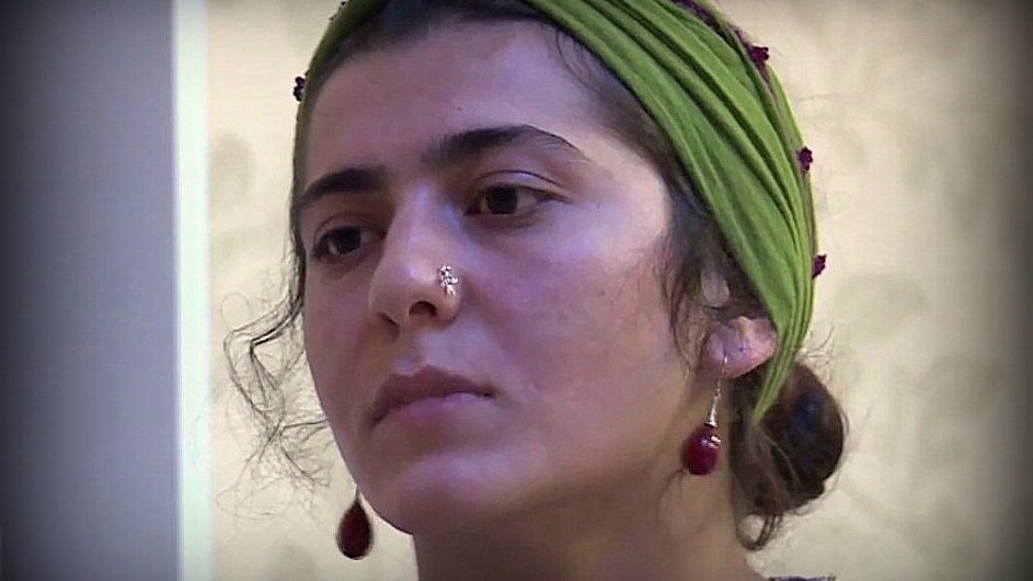 L'image montre une jeune femme avec un regard pensif. Elle porte un foulard vert sur la tête, orné de petites perles. Son visage est en partie encadré par des cheveux bouclés, et elle a un piercing au nez. Elle semble concentrée ou réfléchie, ce qui donne une ambiance contemplative à l'image.