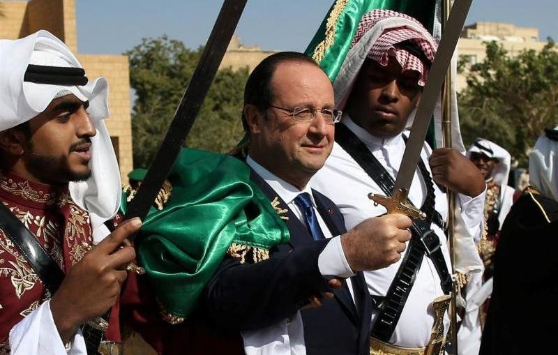L'image montre un homme au centre, portant une veste sombre, accompagné de deux personnes de chaque côté. Ces dernières sont vêtues d'habits traditionnels arabes et tiennent des épées. L'arrière-plan montre un ciel clair et un environnement désertique ou historique, suggérant une ambiance festive ou cérémonielle. L'expression de l'homme central semble joyeuse et accueillante, indiquant qu'il participe à une célébration ou un événement culturel.