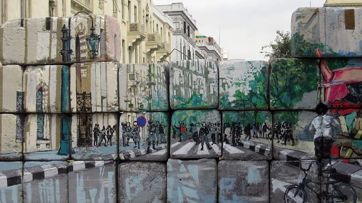 L'image montre un mur de blocs de béton, sur lequel sont peints des paysages urbains. La peinture représente une rue avec des arbres, des piétons et des cyclistes. On peut voir des silhouettes de personnes et des éléments de la ville, comme des lampadaires. Ce contraste entre le mur brut et l'art coloré apporte une touche dynamique à l'environnement urbain. Le ciel est nuageux, ajoutant une atmosphère un peu dramatique à la scène.