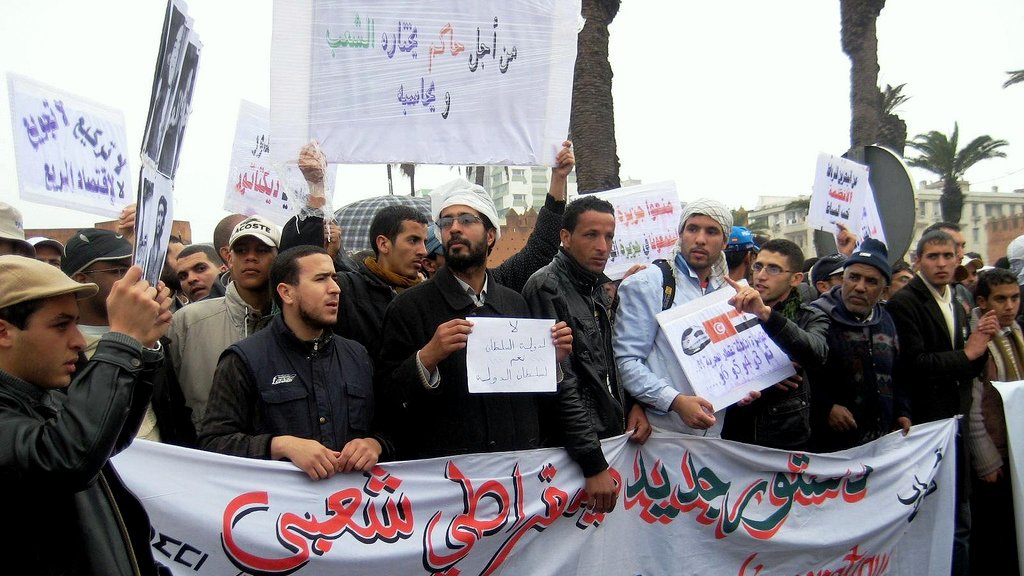 L'image montre un groupe de personnes lors d'une manifestation. Elles tiennent des pancartes et une grande banderole sur laquelle est inscrit en arabe et en français "Pour une constitution démocratique". Les manifestants semblent unis dans leur revendication, et l'environnement montre qu'ils se rassemblent probablement dans une ville, avec des palmiers en arrière-plan. L'atmosphère semble engagée et déterminée.
