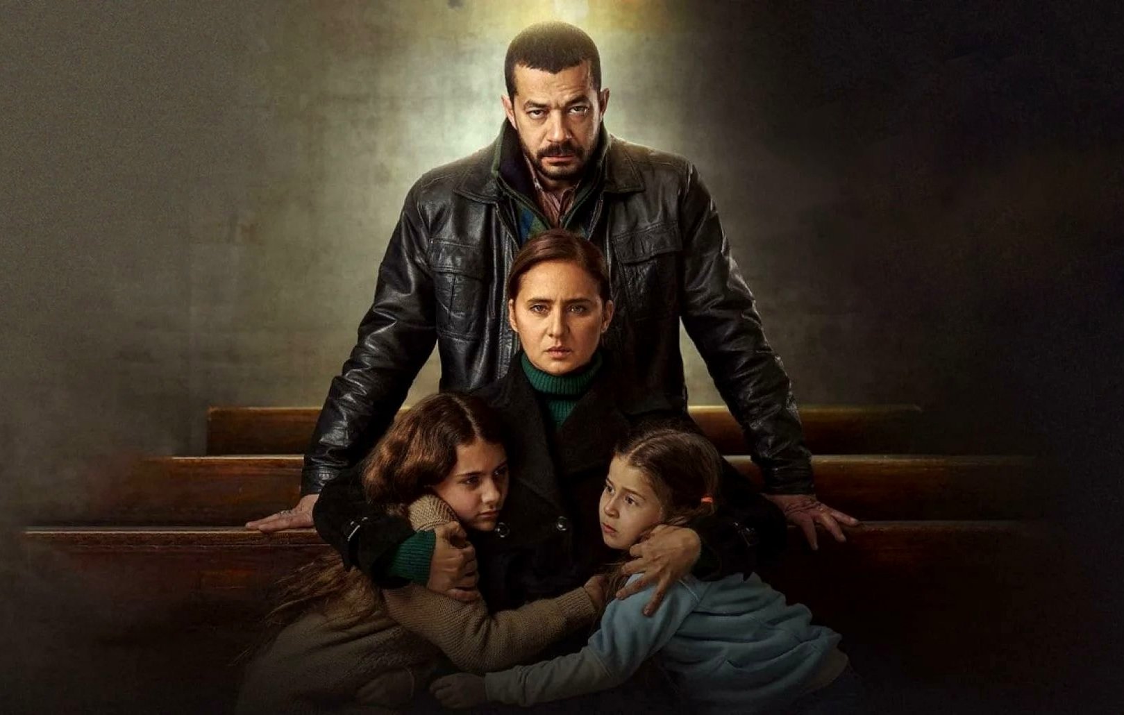 L'image présente une scène dramatique avec quatre personnages. Au centre, une femme est assise, entourée de deux jeunes filles qui semblent chercher protection. La femme a une expression préoccupée, tandis que l'homme derrière elle, vêtu d'une veste en cuir, dégage une présence protectrice et intense. L'éclairage crée un contraste marqué, accentuant l'atmosphère tendue et émotive de la scène. Les éléments de l'environnement, comme les bancs, suggèrent un cadre intérieur, peut-être une église ou un endroit similaire, renforçant le caractère sérieux de la situation.