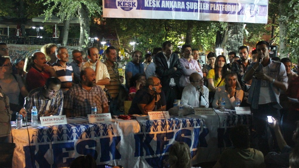 L'image montre une scène de rassemblement ou de meeting en extérieur. Une table est placée au premier plan, où plusieurs personnes sont assises, apparemment des intervenants, pendant qu'une autre personne, debout, s'adresse à l'audience. Un public se tient autour, écoutant attentivement. En arrière-plan, il y a une bannière avec des inscriptions, et l'ambiance semble être celle d'un événement communautaire ou d'une manifestation. Les participants semblent engagés dans une discussion animée.