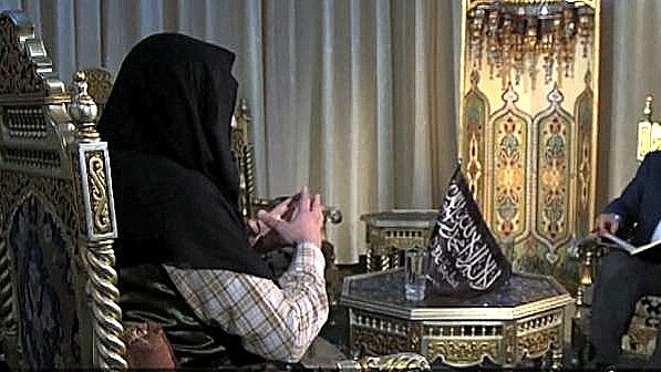 L'image montre deux personnes assises dans un cadre décoratif. L'un des individus porte un hijab et est assis sur un fauteuil orné, tandis que l'autre, en costume, est également assis sur un fauteuil face à lui, tenant un livre. L'environnement semble richement décoré avec des motifs orientaux et des lumières suspendues. L'atmosphère de la scène semble être celle d'une conversation sérieuse.