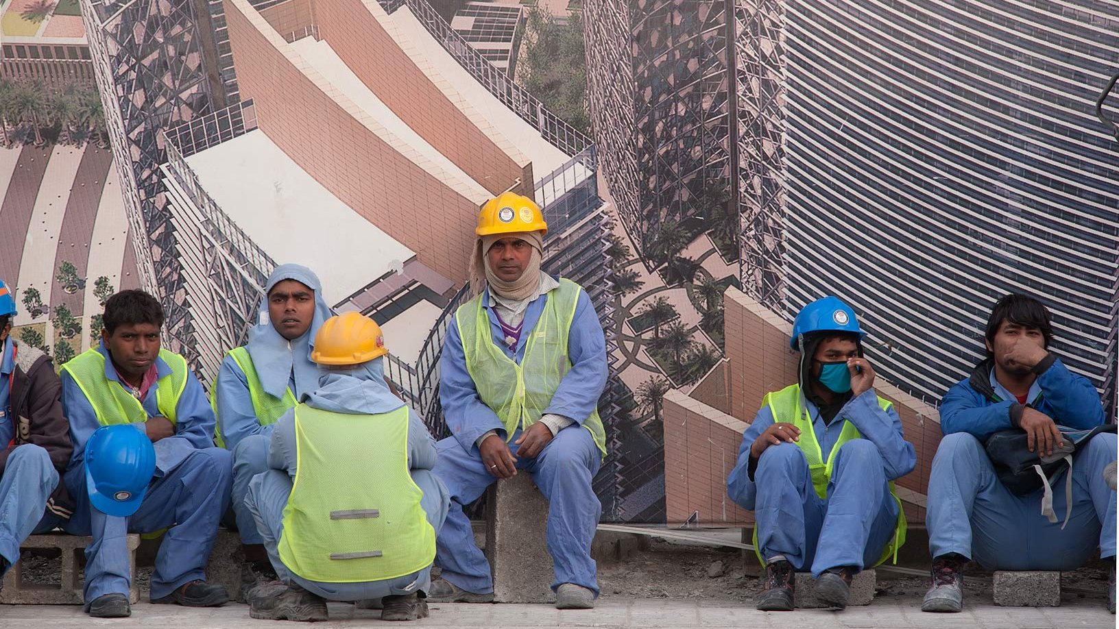 L'image montre un groupe de travailleurs assis sur un mur, probablement en train de faire une pause. Ils portent des vêtements de travail, notamment des gilets de sécurité et des casques de différentes couleurs. L'arrière-plan présente une grande affiche ou un panneau représentant un bâtiment moderne, suggérant qu'ils travaillent sur un site de construction. L'environnement semble urbain et dynamique.