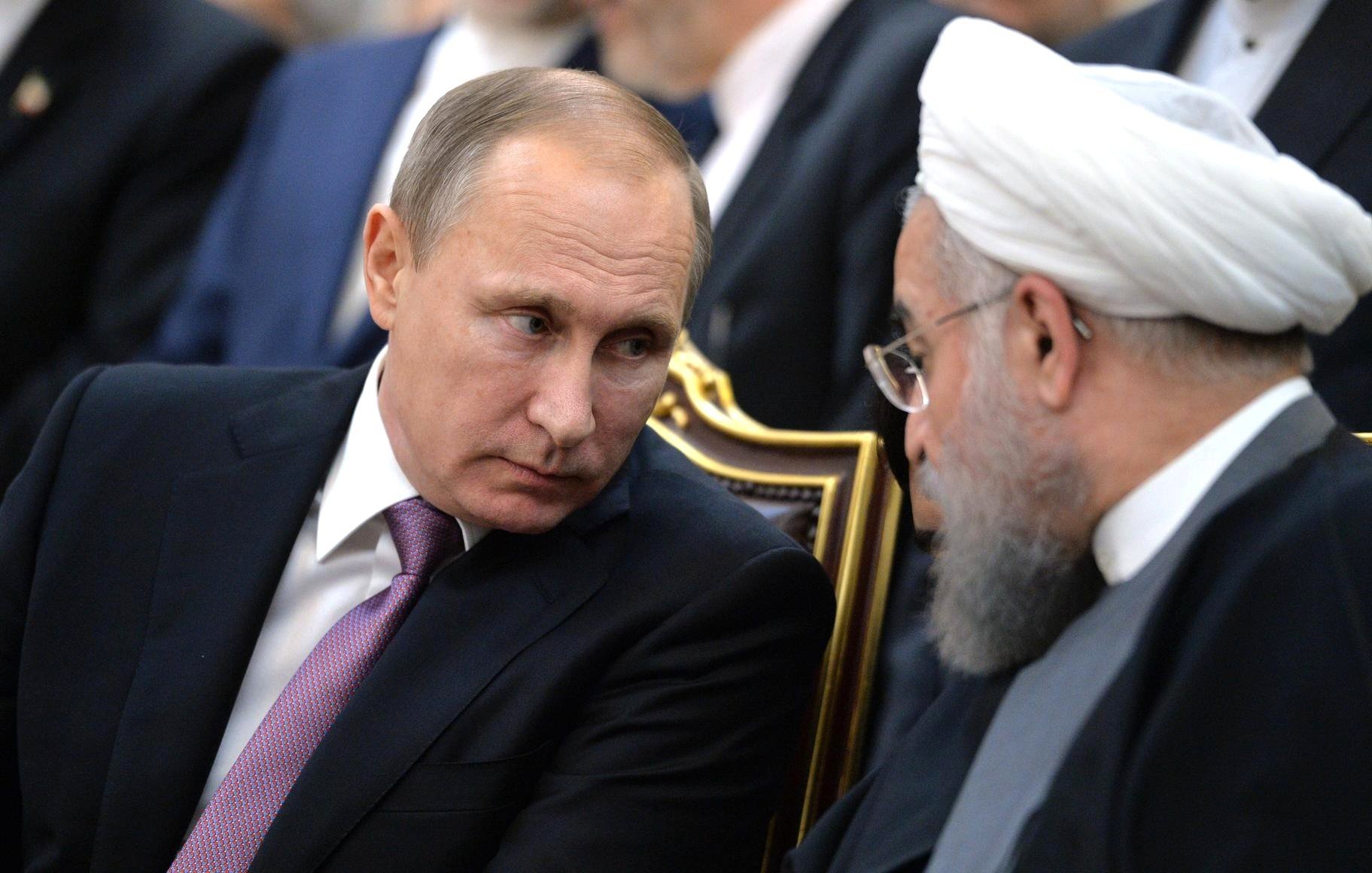 L'image montre deux hommes en costume, assis côte à côte lors d'un événement. L'un d'eux a les cheveux courts et une expression sérieuse, tandis que l'autre porte une barbe grise et semble en train de discuter. Ils sont entourés d'autres personnes floues en arrière-plan. Leur posture suggère une conversation attentive et concentrée.