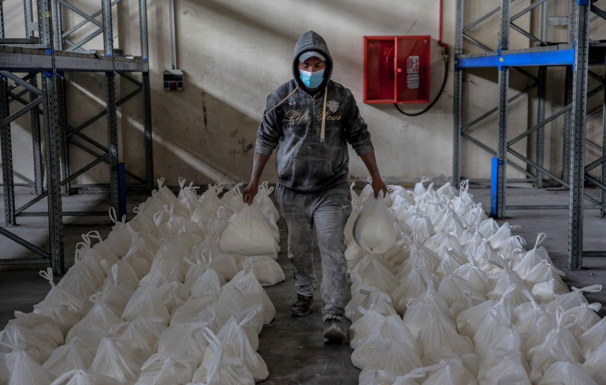Sur cette image, on voit une personne portant un sweat à capuche gris et un masque de protection. Elle marche à travers une pièce de stockage ou un entrepôt, tenant deux sacs blancs en plastique. De nombreux sacs similaires sont disposés de manière ordonnée sur le sol autour de la personne. Les murs de la salle semblent ternes et l'éclairage suggère un environnement de travail modeste. Au fond, on peut voir une boîte de protection contre les incendies fixée au mur.