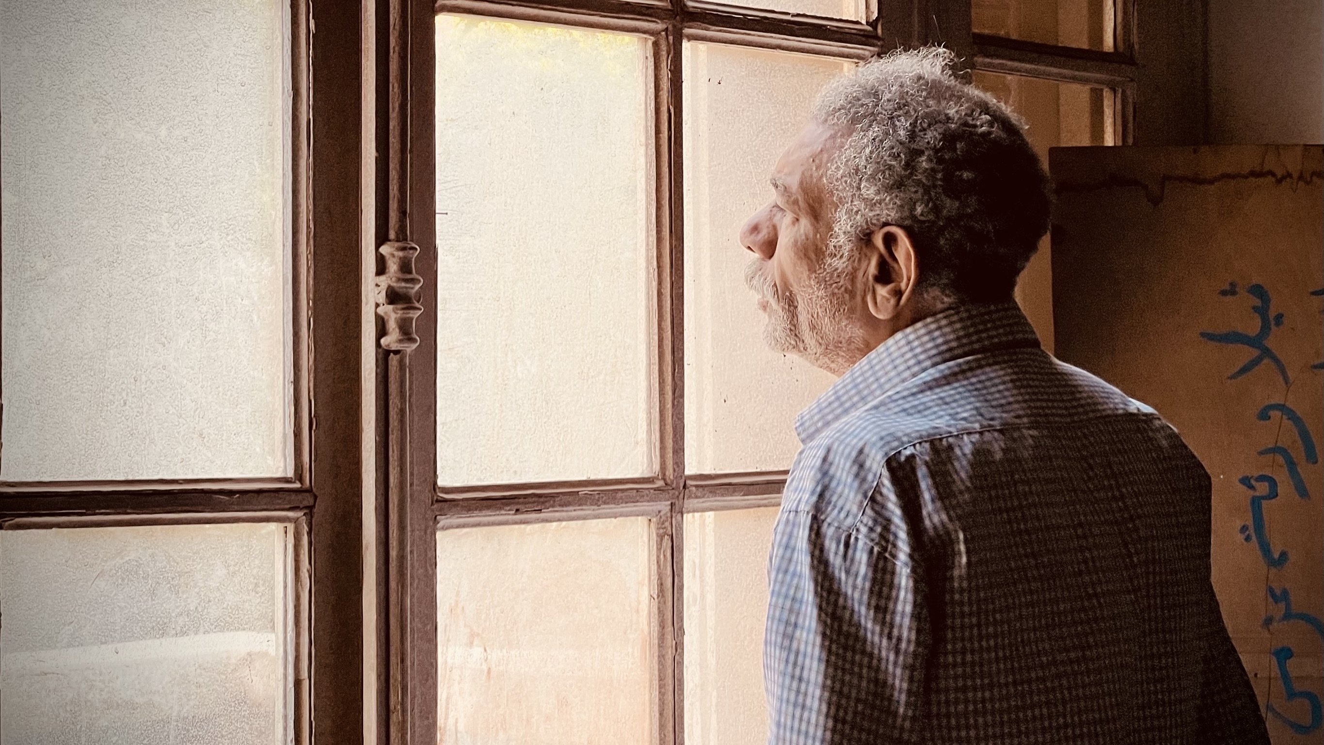 L'image montre un homme âgé qui semble contempler à travers une fenêtre. Il est habillé d'une chemise à motifs et a une posture pensive. La lumière naturalisée entre par la fenêtre, illuminant doucement son visage et créant une ambiance calme. On peut aussi apercevoir des murs en arrière-plan, ajoutant à l'atmosphère de réflexion du moment.