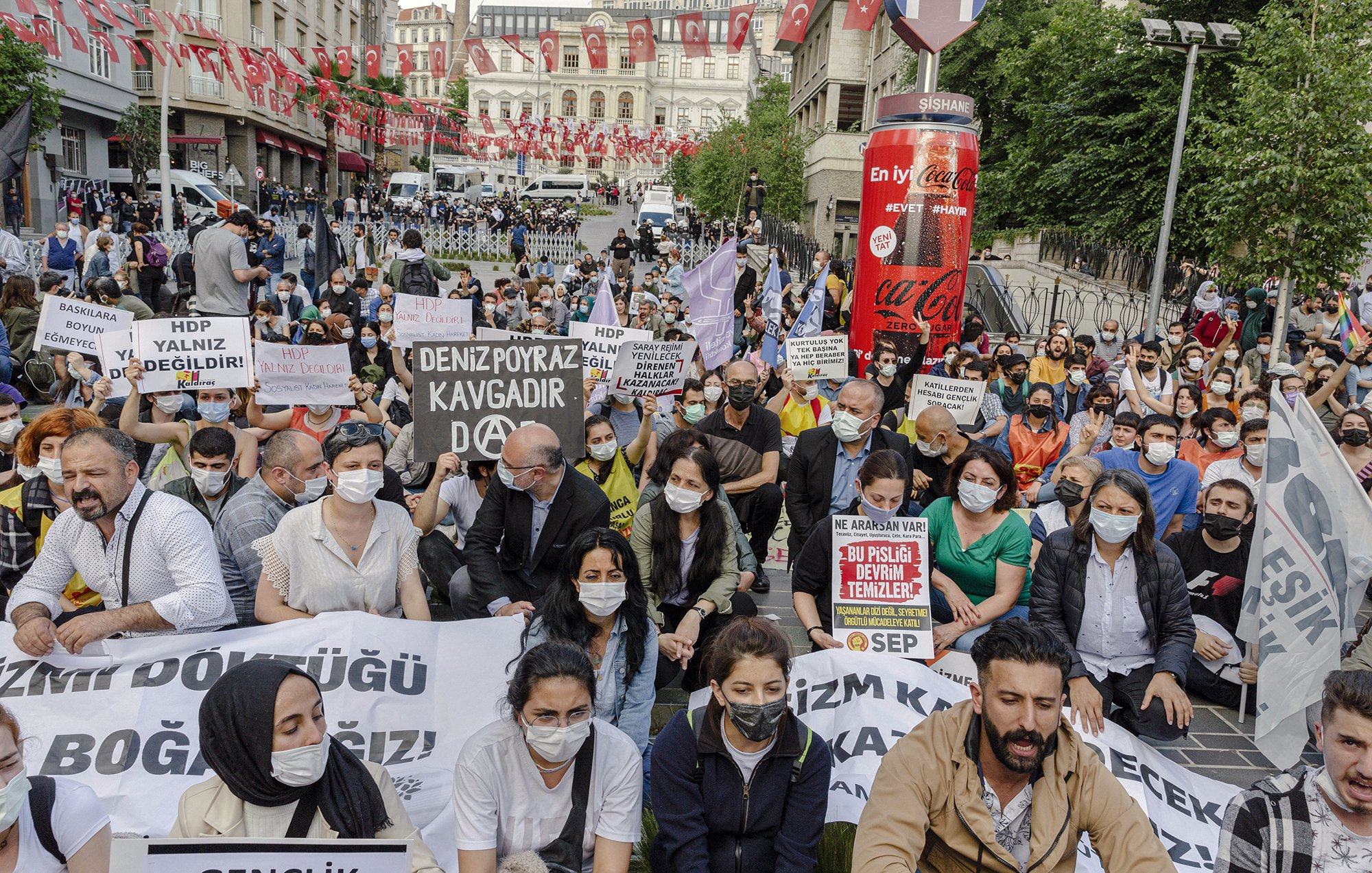 L'image montre une manifestation en plein air avec de nombreuses personnes rassemblées dans une rue. Les manifestants portent des masques et brandissent des pancartes avec des slogans. L'atmosphère semble mobilisée, et il y a beaucoup de drapeaux en arrière-plan. Les participants sont assis pour exprimer leur message, signalant une volonté de protester pacifiquement.