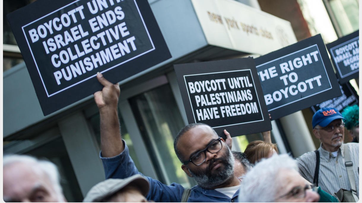 L'image montre une manifestation avec des personnes tenant des pancartes. Les pancartes portent des messages appelant au boycott jusqu'à ce qu'Israël mette fin à des mesures de punition collective et jusqu'à ce que les Palestiniens aient la liberté. On peut voir des participants de différents âges, tous unis par des revendications similaires. L'arrière-plan indique un environnement urbain avec des bâtiments.