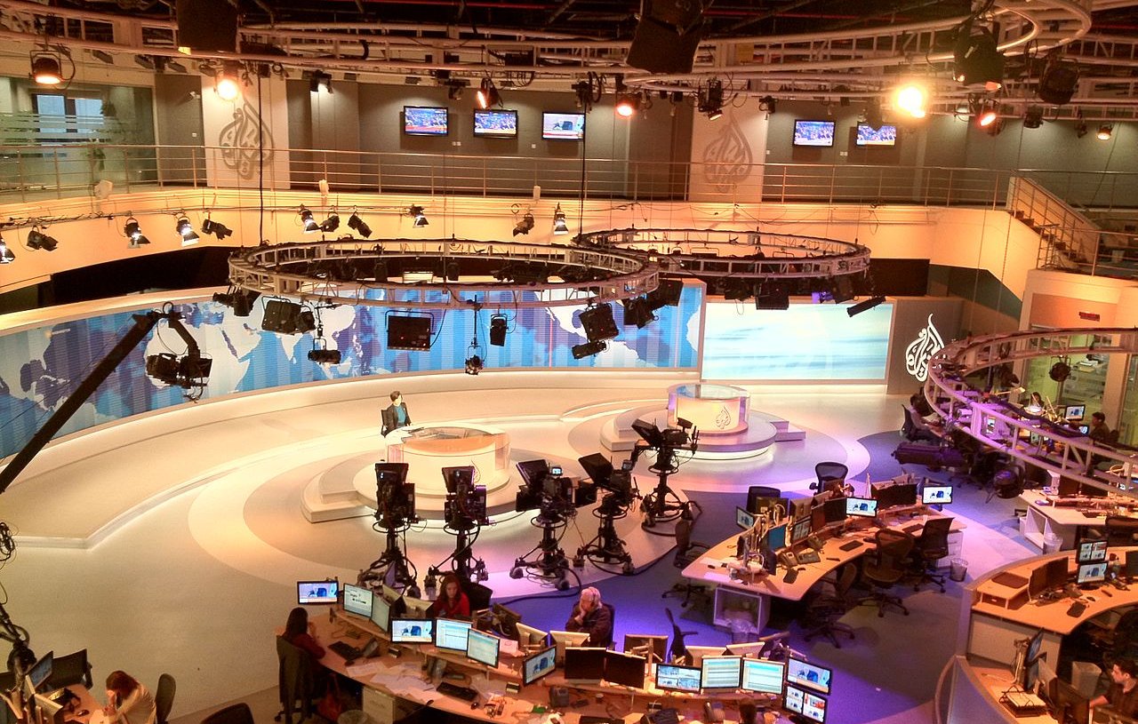 L'image montre un studio de télévision moderne, probablement utilisé pour l'information. On y voit plusieurs caméras placées sur des trépieds, ainsi que des moniteurs et des bureaux avec des ordinateurs. Le décor est élégant, avec des panneaux de fond aux couleurs douces et des éclairages suspendus. Au centre, il y a un espace pour le présentateur, entouré d'une atmosphère professionnelle et technologique. L'ensemble donne une impression de dynamisme et de préparation pour diffuser des nouvelles en direct.