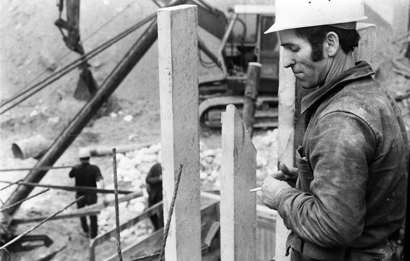 L'image montre un homme en train de travailler sur un chantier de construction. Il porte un casque de protection blanc et semble concentré sur sa tâche. À proximité, on peut voir des éléments en béton vertical, probablement des murs ou des supports. En arrière-plan, des ouvriers sont également visibles, et une machine lourde est à l'œuvre. L'image est en noir et blanc, ce qui lui donne un aspect rétro et saisissant. L'ambiance générale évoque le travail manuel et l'effort dans le domaine de la construction.