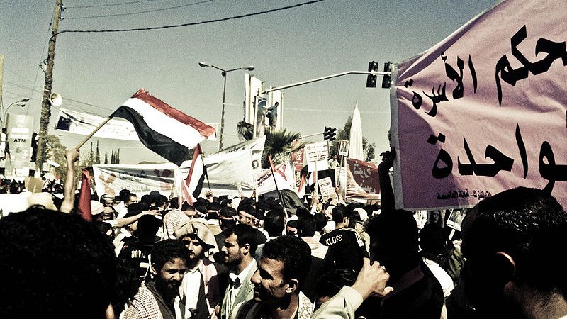 L'image montre une manifestation animée avec une grande foule de personnes. Des drapeaux flottent dans l'air, en particulier un drapeau qui semble être celui du Yémen. On peut voir des hommes et des femmes rassemblés avec des expressions déterminées. Des pancartes sont tenues par des manifestants, certaines comportant des inscriptions en arabe. L'atmosphère est dynamique, reflétant le désir de changement ou de revendication au sein de la foule. Les bâtiments environnants et les lignes électriques en arrière-plan ajoutent un contexte urbain à la scène.