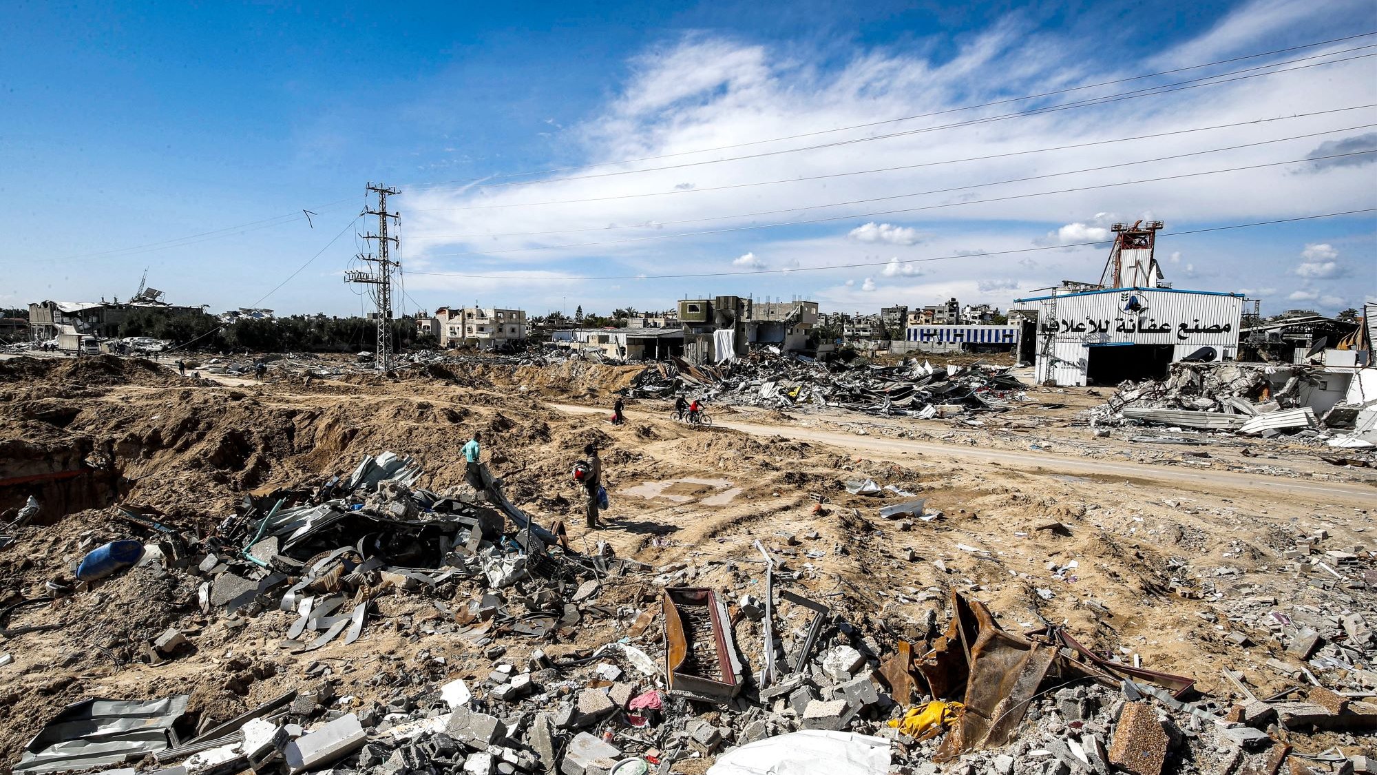 L'image montre un paysage dévasté, probablement dans une zone de conflit ou après une catastrophe. Le sol est jonché de débris, avec des morceaux de béton et de métal éparpillés. On peut apercevoir quelques personnes se déplaçant parmi les ruines, tandis qu'à l'arrière-plan, des bâtiments endommagés se dressent sous un ciel partiellement nuageux. Les lignes électriques sont visibles, indiquant une infrastructure perturbée. L'ambiance apparaît sombre et désolée, témoignant de la destruction qui a eu lieu.