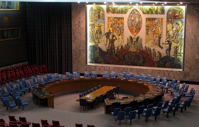L'image montre l'intérieur d'une salle de réunion, probablement celle du Conseil de sécurité des Nations Unies. On y voit un grand panneau mural coloré représentant des scènes symboliques. La salle est circulaire avec une table ovale au centre, entourée de chaises disposées en arcs. Les chaises sont de différentes couleurs, majoritairement bleues et rouges. L'éclairage est doux, mettant en valeur les œuvres d'art sur le mur. L'atmosphère semble formelle et solennelle, propice aux discussions diplomatiques.
