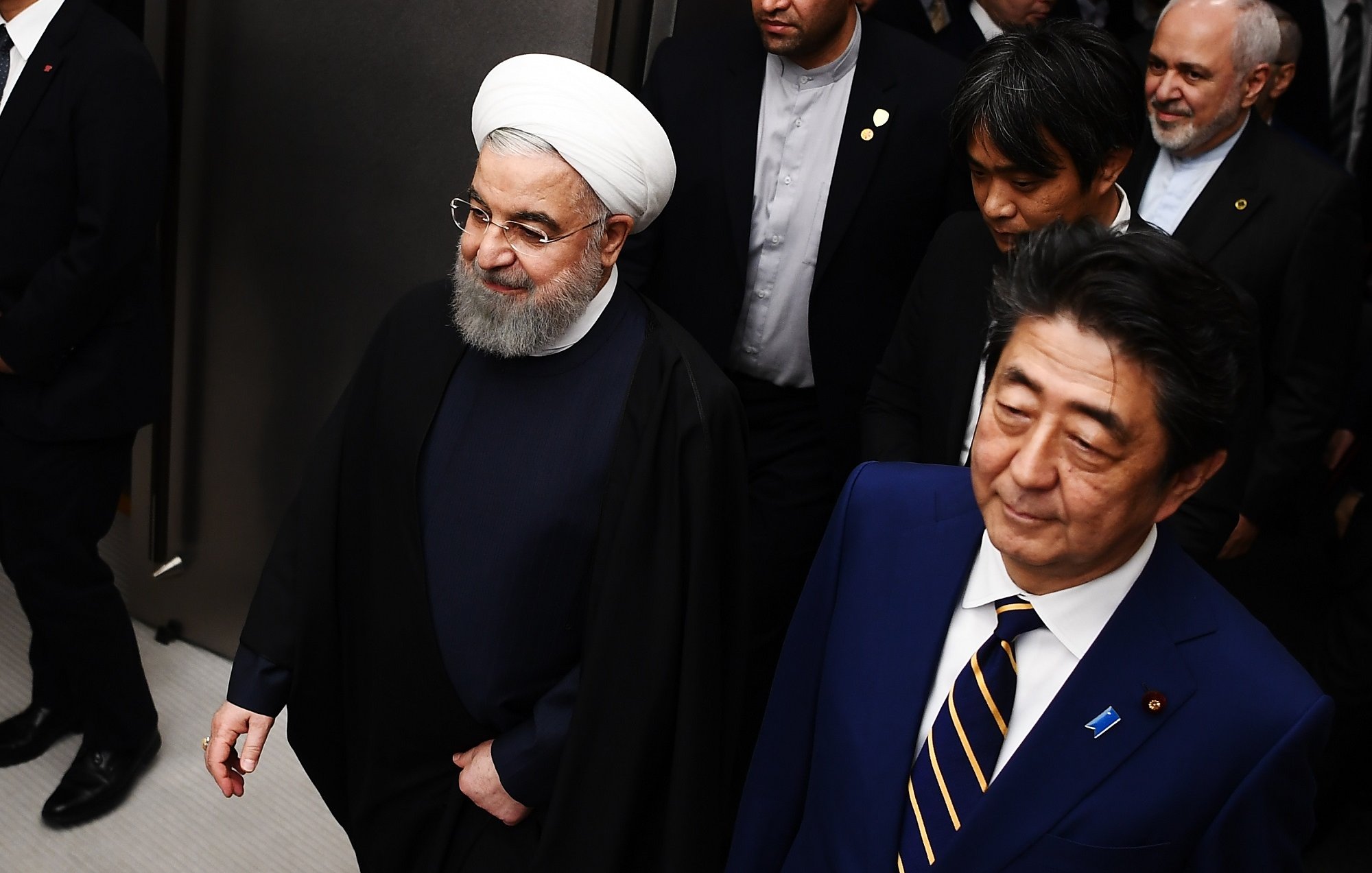 L'image montre deux hommes marchant côte à côte dans un environnement formel. L'un d'eux porte une tenue sombre avec une coiffure distinctive et un turban blanc, tandis que l'autre est habillé de manière formelle en costume. Ils semblent être entourés de plusieurs autres personnes habillées aussi de manière professionnelle. L'atmosphère dégage un air de réunion ou d'événement officiel.