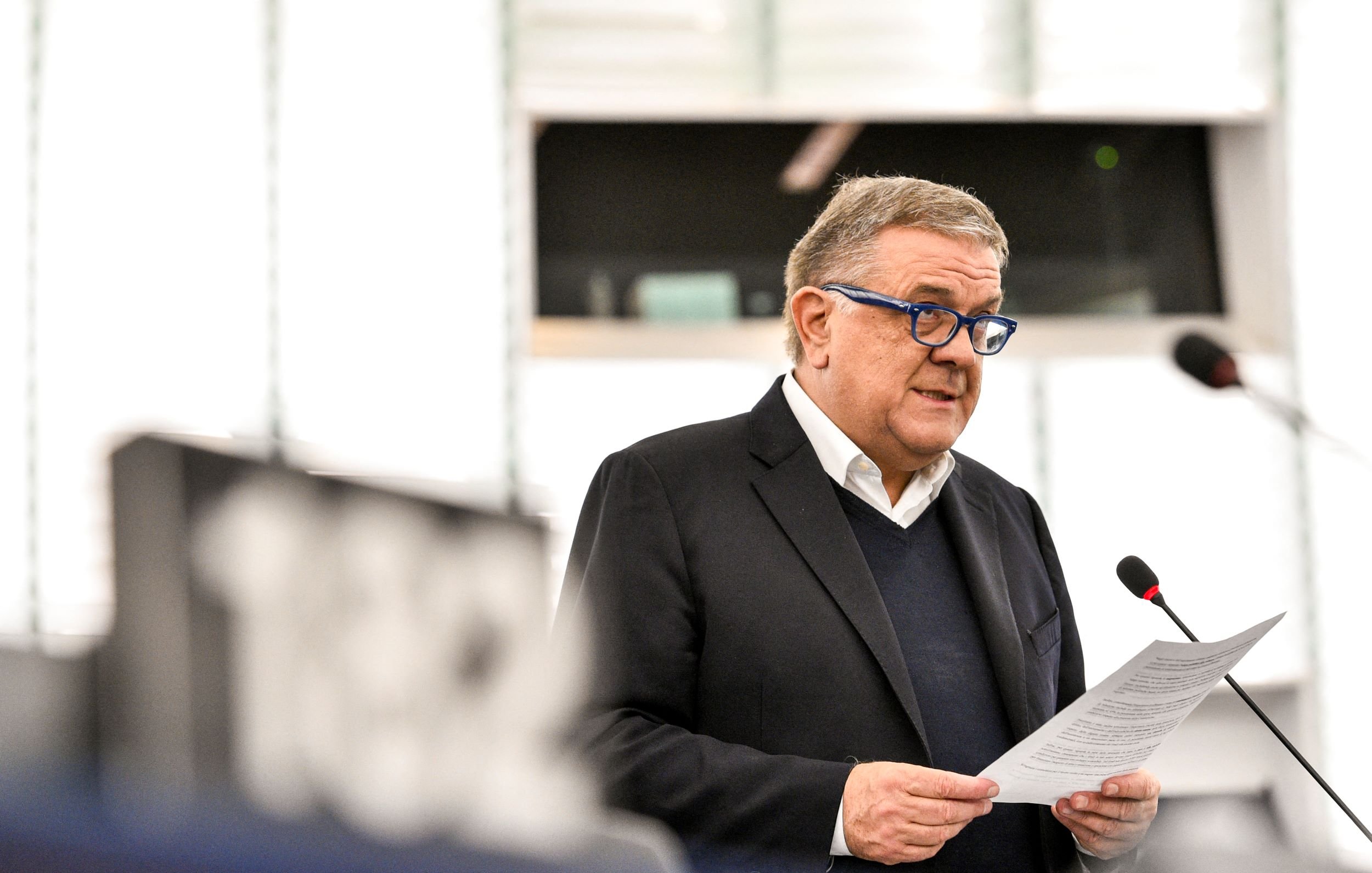 L'image montre un homme qui parle au micro lors d'une session au Parlement. Il porte des lunettes bleues et un costume sombre. Il semble lire un discours ou un document. L'environnement est celui d'une salle de réunion institutionnelle, avec un éclairage moderne et des panneaux en arrière-plan.