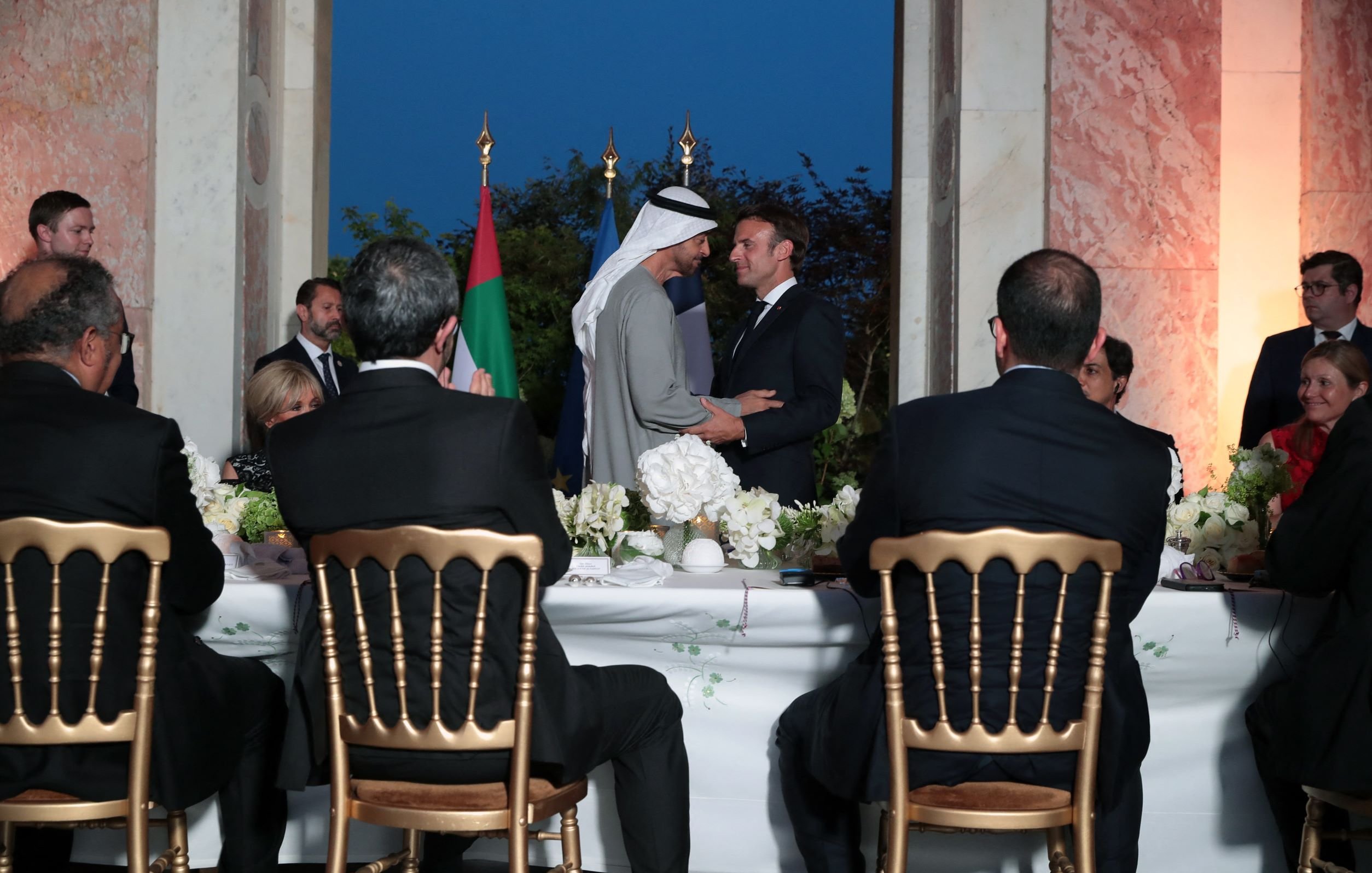 L'image montre une scène formelle dans un cadre élégant, probablement lors d'un dîner ou d'une réception. Au centre, deux hommes se serrent la main. On peut voir des drapeaux en arrière-plan, suggérant un contexte diplomatique. À la table, plusieurs personnes assistent à l'événement, avec une attention particulière sur les deux protagonistes. L'ambiance est chaleureuse et conviviale, marquée par des éléments de décoration soignés, comme des fleurs.