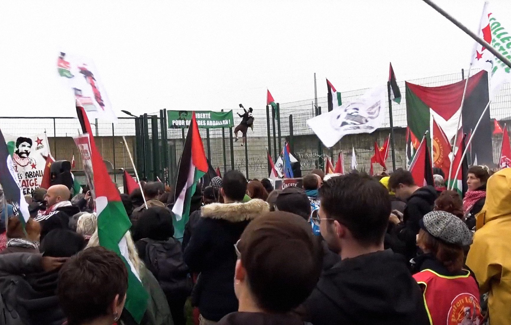 L'image montre une foule rassemblée lors d'une manifestation. Des personnes tiennent des drapeaux de différentes couleurs, dont des drapeaux palestiniens et d'autres symboles politiques. On peut voir une grande structure en arrière-plan, probablement une barrière ou une clôture, avec des bannières accrochées. L'atmosphère semble dynamique et engagée, reflétant une ambiance de protestation ou de revendication. Le temps paraît nuageux, ajoutant à l'intensité de l'événement.