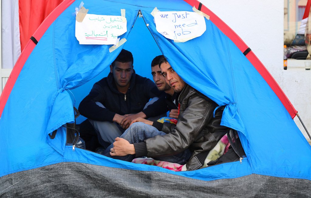 L'image montre un groupe de trois jeunes hommes assis à l'intérieur d'une tente. Ils semblent discuter ensemble, et des pancartes en arabe et en anglais sont visibles à l'extérieur de la tente, indiquant "Nous avons juste besoin d'un emploi". La tente est de couleur bleue, et l'ambiance générale évoque un sentiment de rassemblement autour d'une cause.