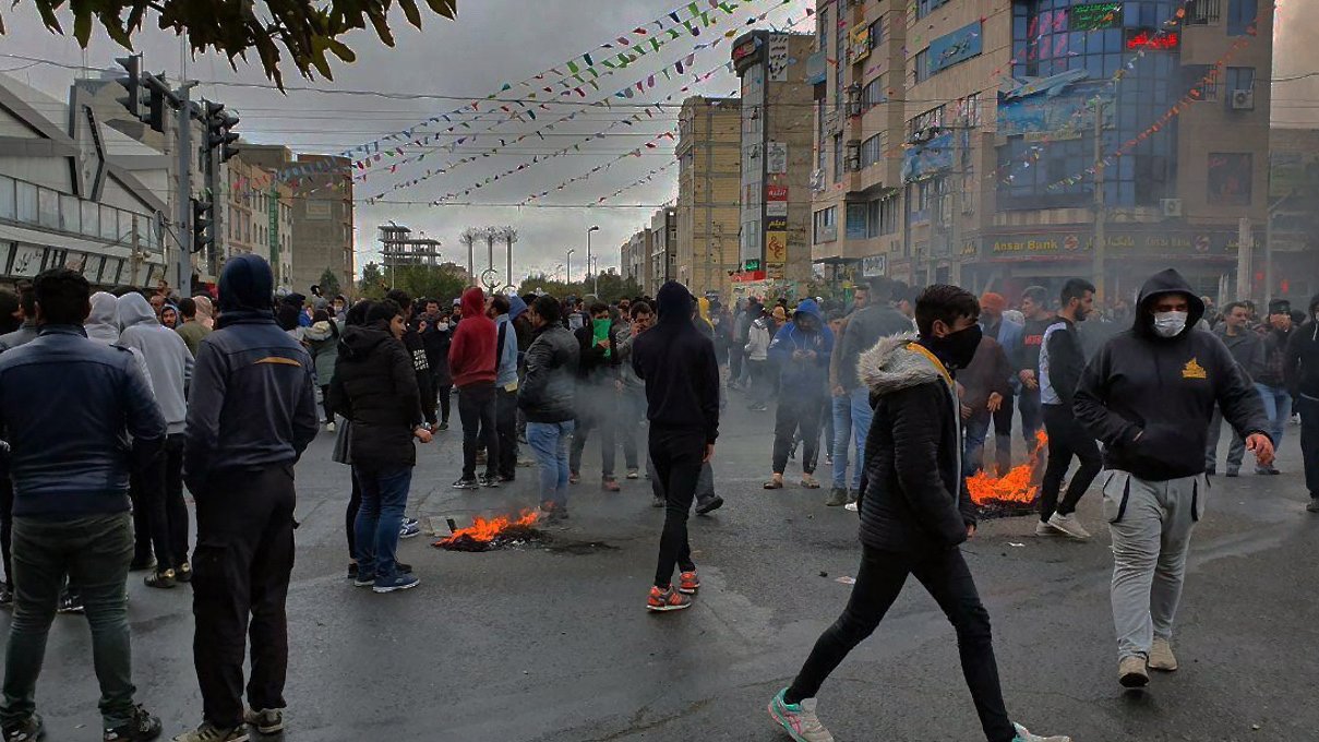 L'image montre une foule rassemblée dans une rue, probablement lors d'une manifestation. Plusieurs personnes portent des vêtements chauds et des masques. On peut observer des petits feux sur la chaussée, entourés de fumée. L'atmosphère semble tendue, avec des bâtiments et des décorations en arrière-plan. Le ciel est nuageux, ajoutant à l'ambiance sombre de la scène.
