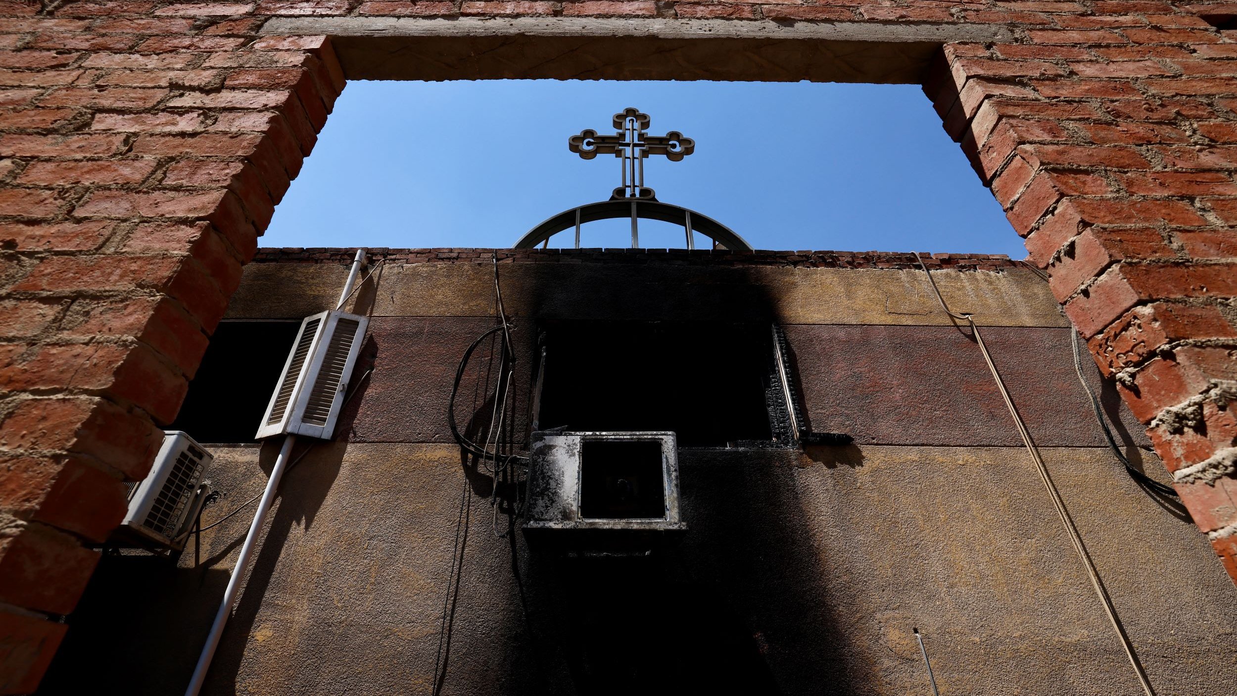 L'image montre un cadre en briques laissant entrevoir un ciel bleu, avec une croix noire au sommet d'une structure. On peut également voir des éléments d'électricité sur le mur, et une partie du mur semble avoir subi des dommages dus à un incendie, avec des marques noires. L'ensemble dégage une atmosphère de contraste entre la lumière et l'ombre, ainsi qu'une impression de désolation.