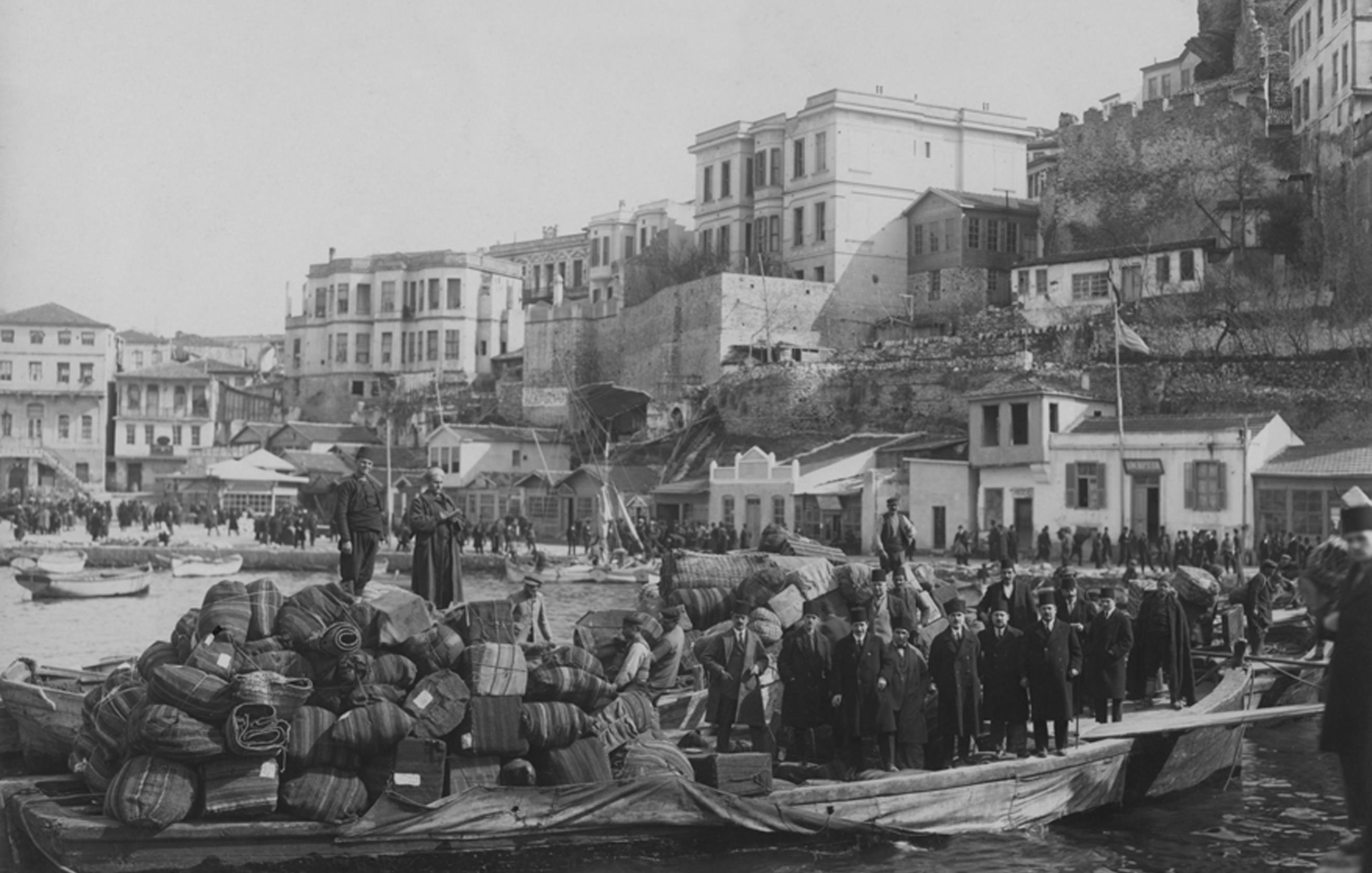 Cette image en noir et blanc montre une scène portuaire historique. On peut voir une barge chargée de marchandises, notamment des fûts et des sacs, avec des personnes debout dessus. En arrière-plan, il y a des bâtiments qui semblent faire partie d'une ville côtière, avec des structures en pierre et des maisons aux balcons. La côte est animée, avec d'autres bateaux et des gens sur le rivage, créant une atmosphère de commerce maritime. La lumière et le contraste évoquent une ambiance d'époque.