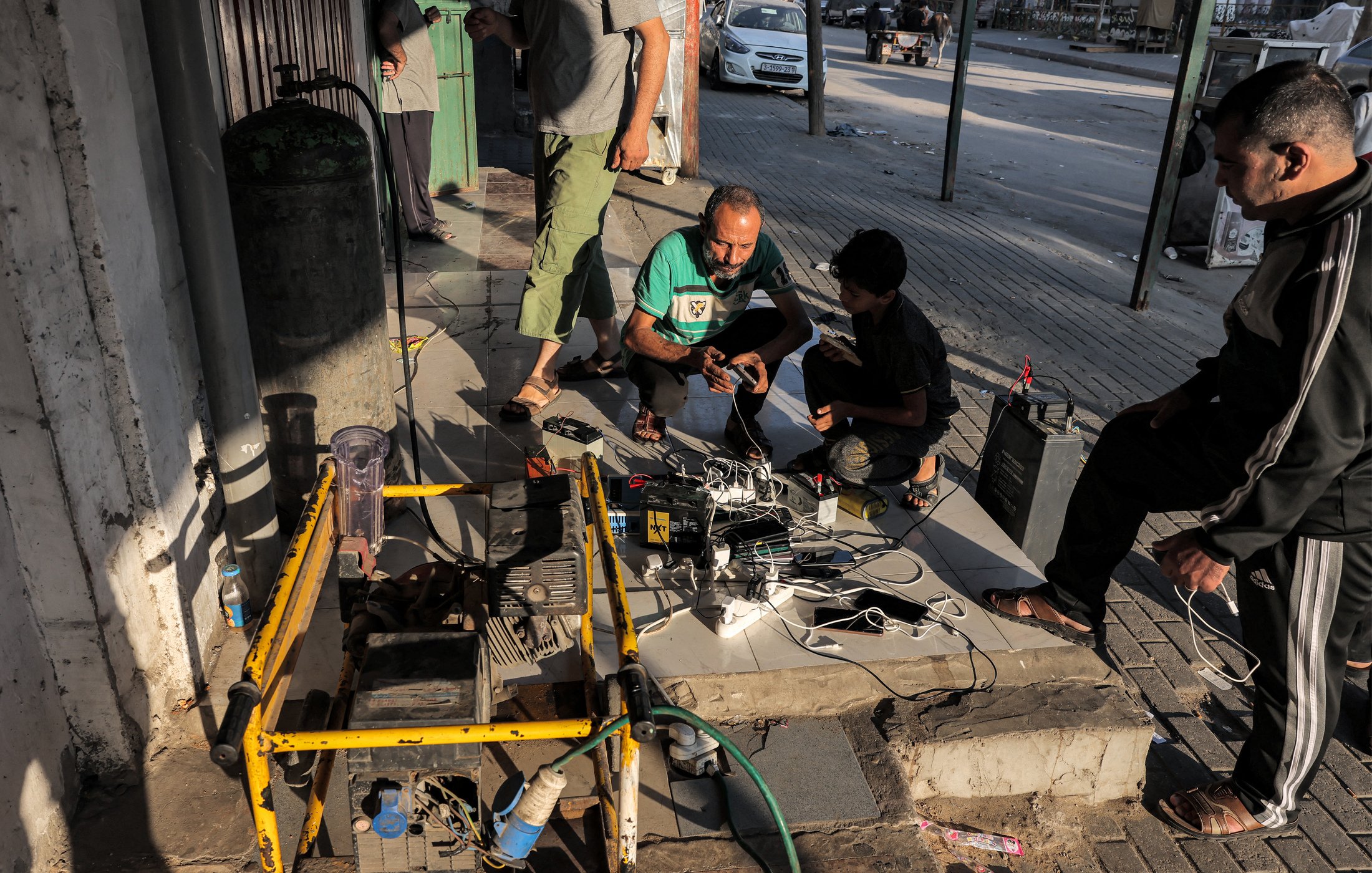 Cette image montre un groupe de personnes, principalement des hommes, rassemblées autour d'un générateur et de plusieurs chargeurs de téléphone portable. Ils se trouvent à l'extérieur, probablement sur un trottoir ou une zone piétonne pavée. Plusieurs téléphones portables sont connectés aux chargeurs qui sont branchés au générateur. À l'arrière-plan, on peut voir une voiture garée et des bâtiments. L'atmosphère semble indiquer une situation où les gens utilisent un générateur pour charger leurs appareils en l'absence d'une source d'électricité régulière.